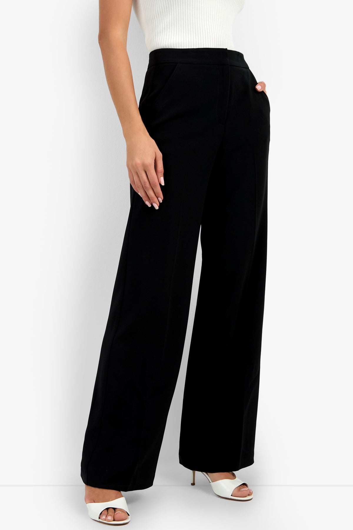 A woman wears the Sadie Pant—high-waisted, wide-leg black pants—paired with a sleeveless white top and white open-toe heels, standing with one hand in her pocket against a plain background.