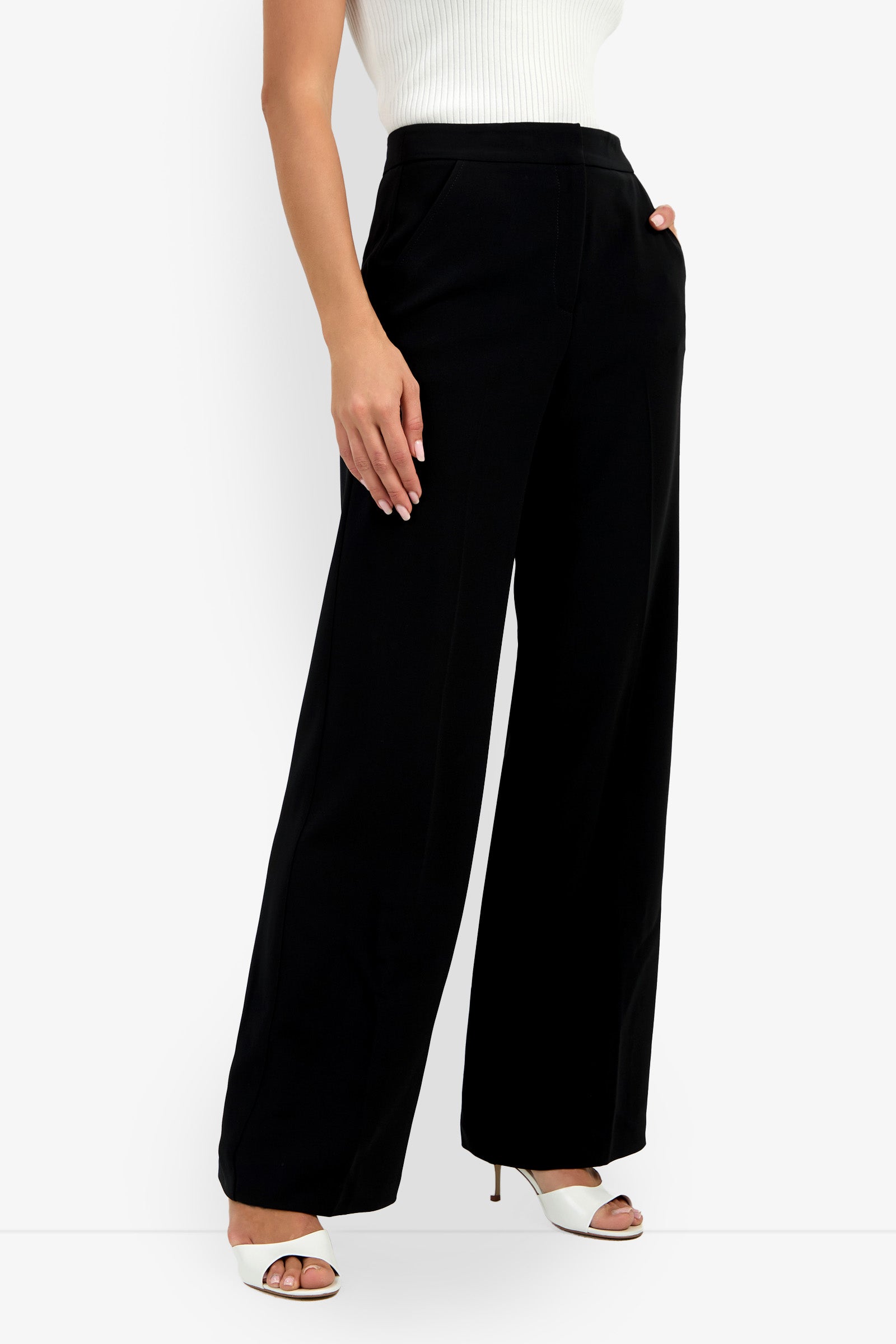 A person wearing a white sleeveless ribbed top, Sadie Pant in high-waisted black wide-leg style, and white open-toe heeled sandals stands with one hand in their pocket against a plain background.