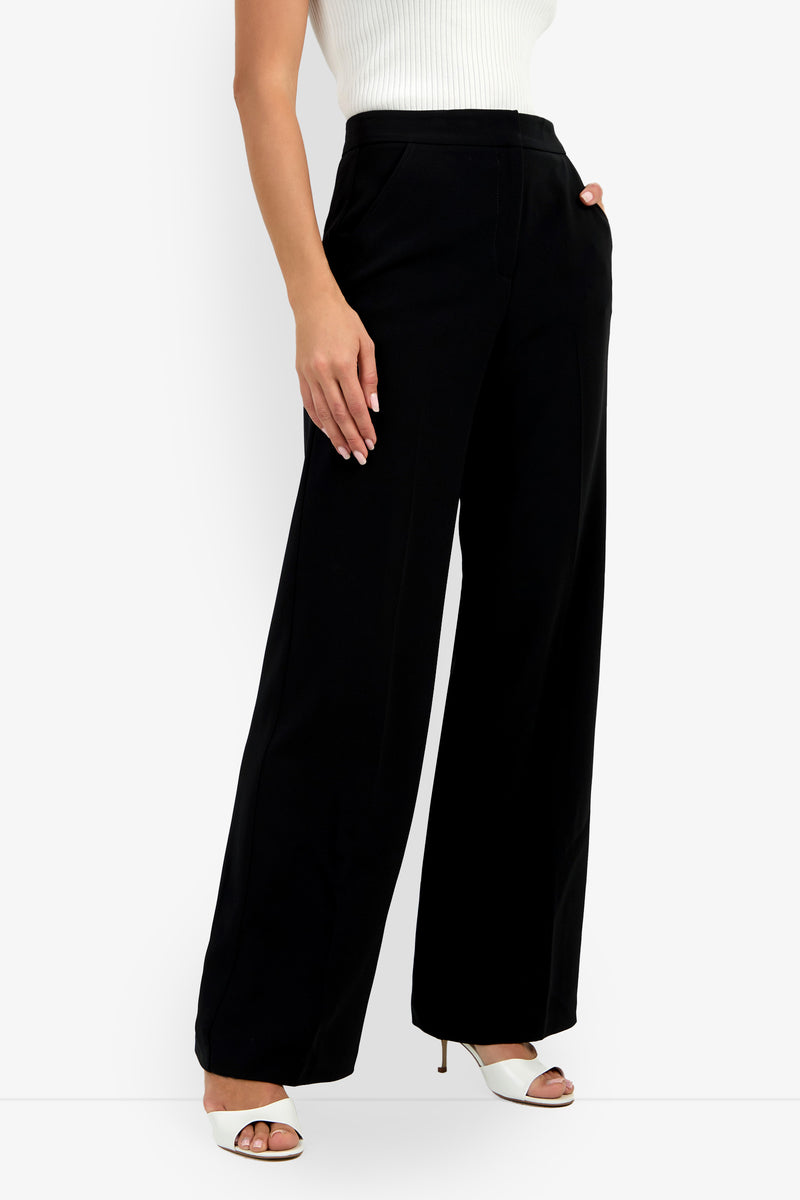 A person wearing a white sleeveless ribbed top, Sadie Pant in high-waisted black wide-leg style, and white open-toe heeled sandals stands with one hand in their pocket against a plain background.