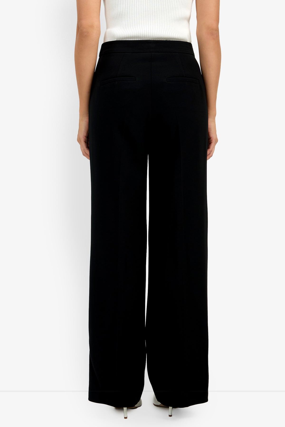 A person stands with their back to the camera against a white background, wearing the Sadie Pant—high-waisted, wide-leg black trousers with two back welt pockets—and a white sleeveless top.