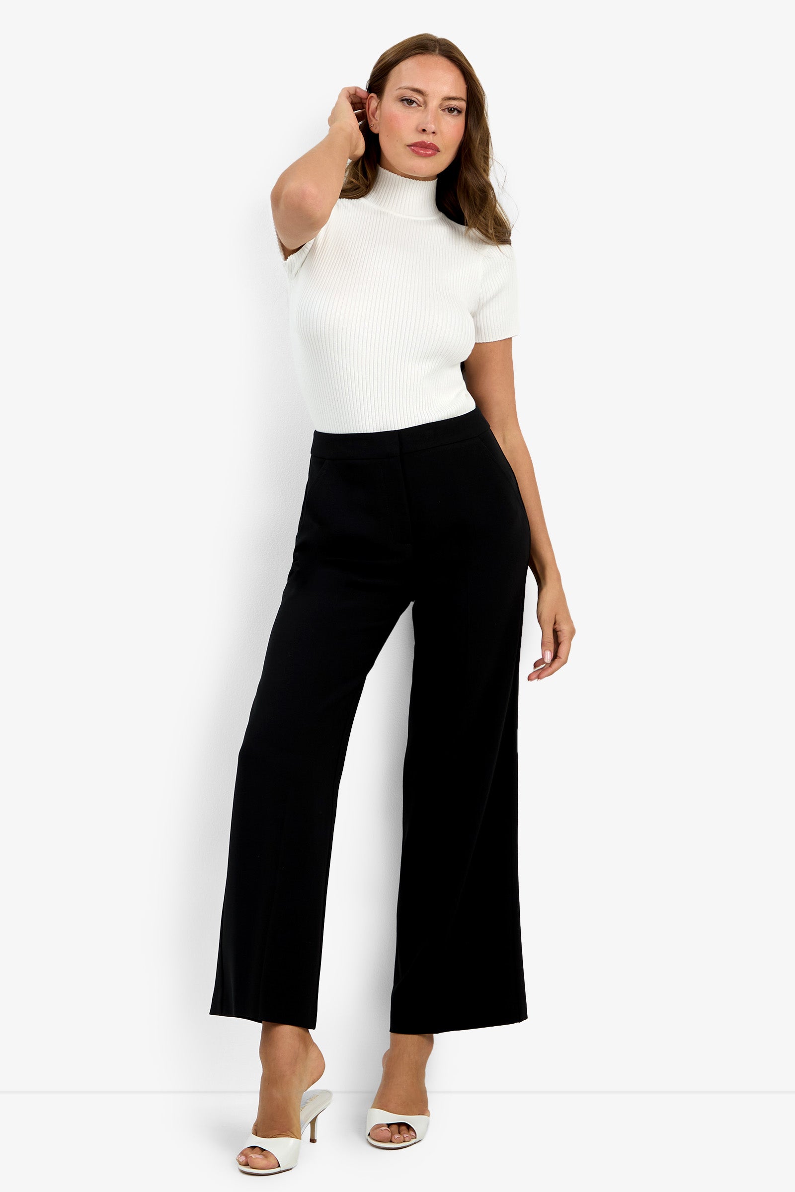 A woman poses against a white background wearing high-waisted Sadie Petite Pants in black, a fitted white short-sleeve turtleneck top, and white heeled sandals. She touches her hair with one hand and has a neutral expression.