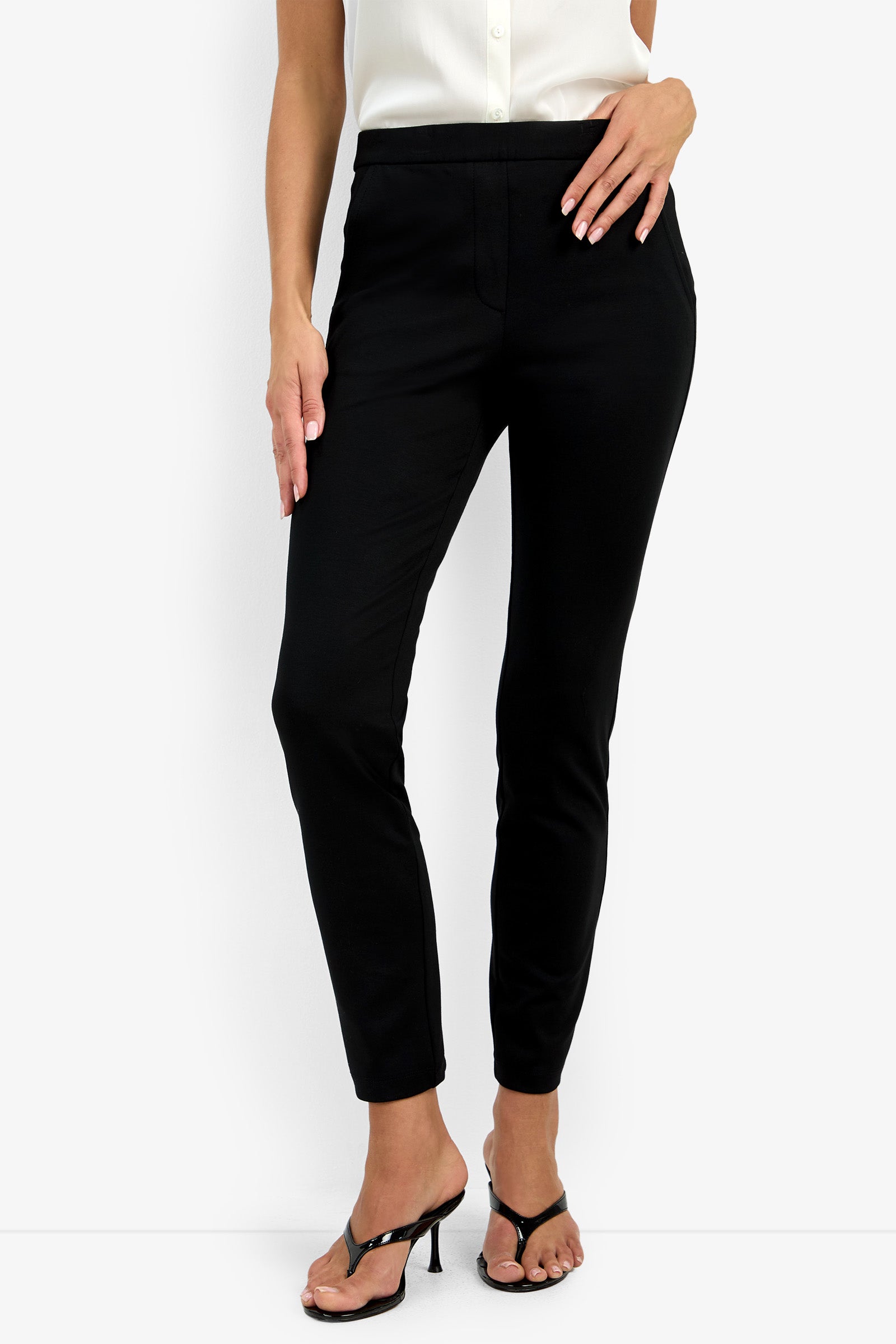 A woman models the Honey Pant—slim-fit black pants—paired with a sleeveless white blouse and black heeled sandals, posing with one hand on her hip against a plain white background.