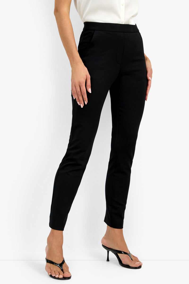 A person wearing the Honey Pant in black slim-fit style, paired with a white blouse and black heeled sandals, stands with one leg slightly forward against a plain white background.