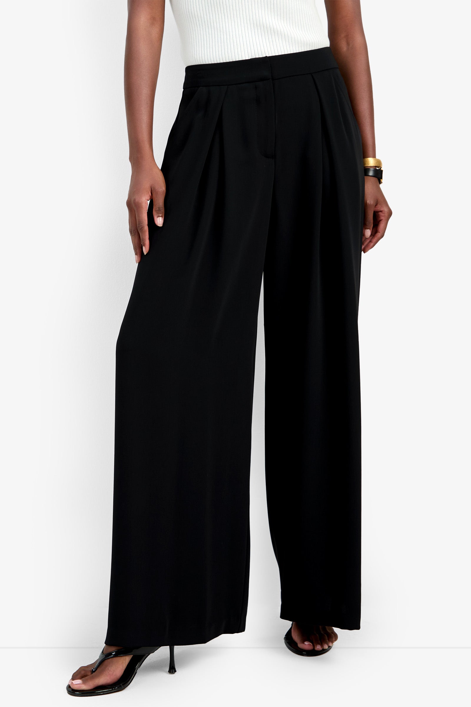 A person wearing a white sleeveless top, the black Saige Pant, black high-heeled sandals, and a gold bangle stands against a white background.