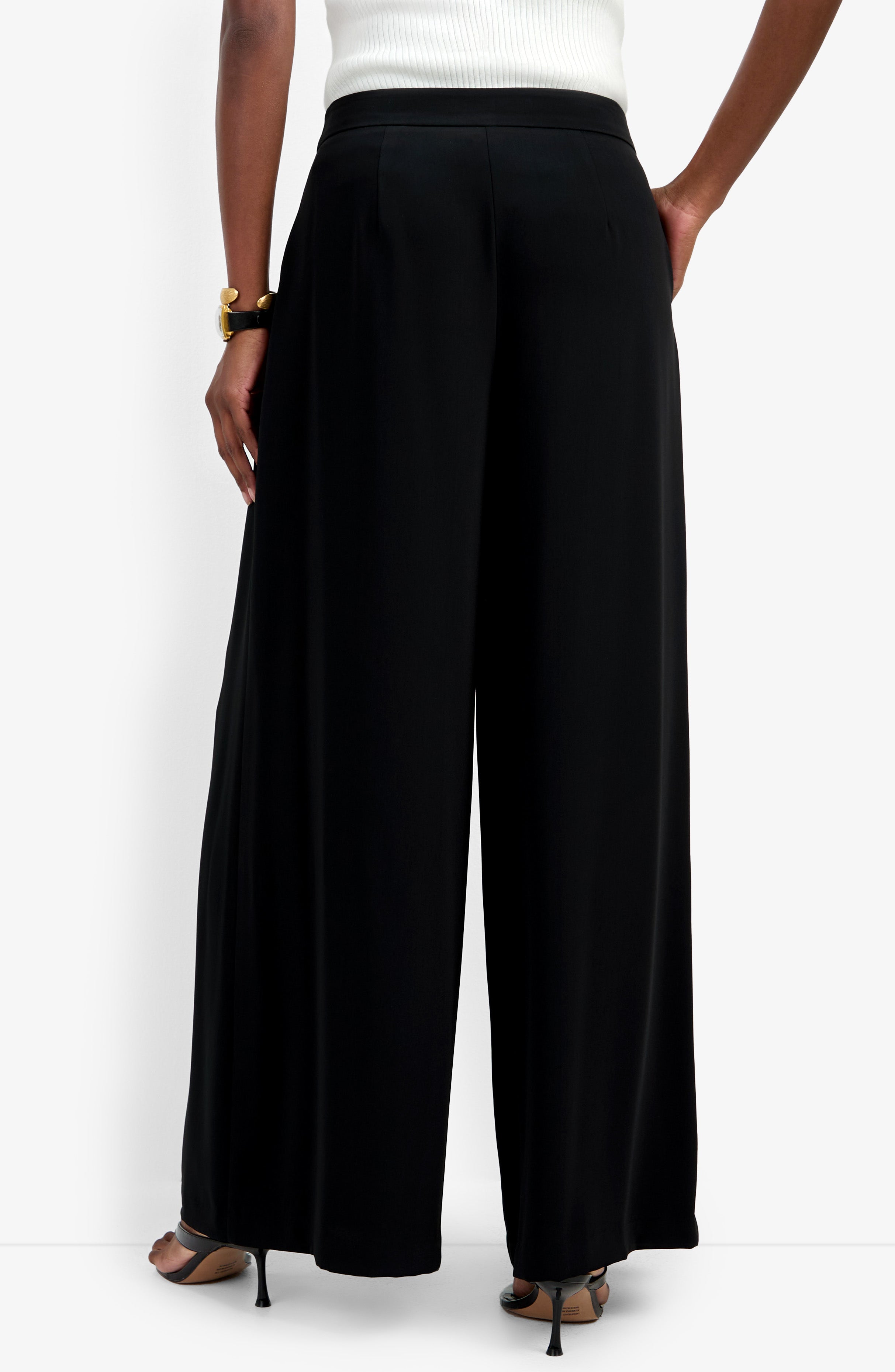 A person wearing the Saige Pant in black, a white top, black heeled sandals, and a gold bracelet stands with one hand in their pocket, photographed from the back against a plain white background.