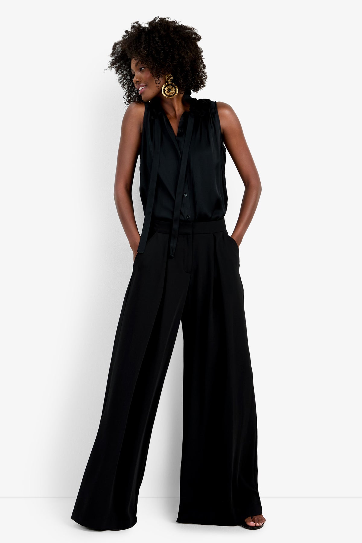 A woman with curly hair wears wide-leg black Saige Long Pants and a sleeveless black blouse with a tie detail. She stands against a plain white background, hands in pockets, looking to the side.
