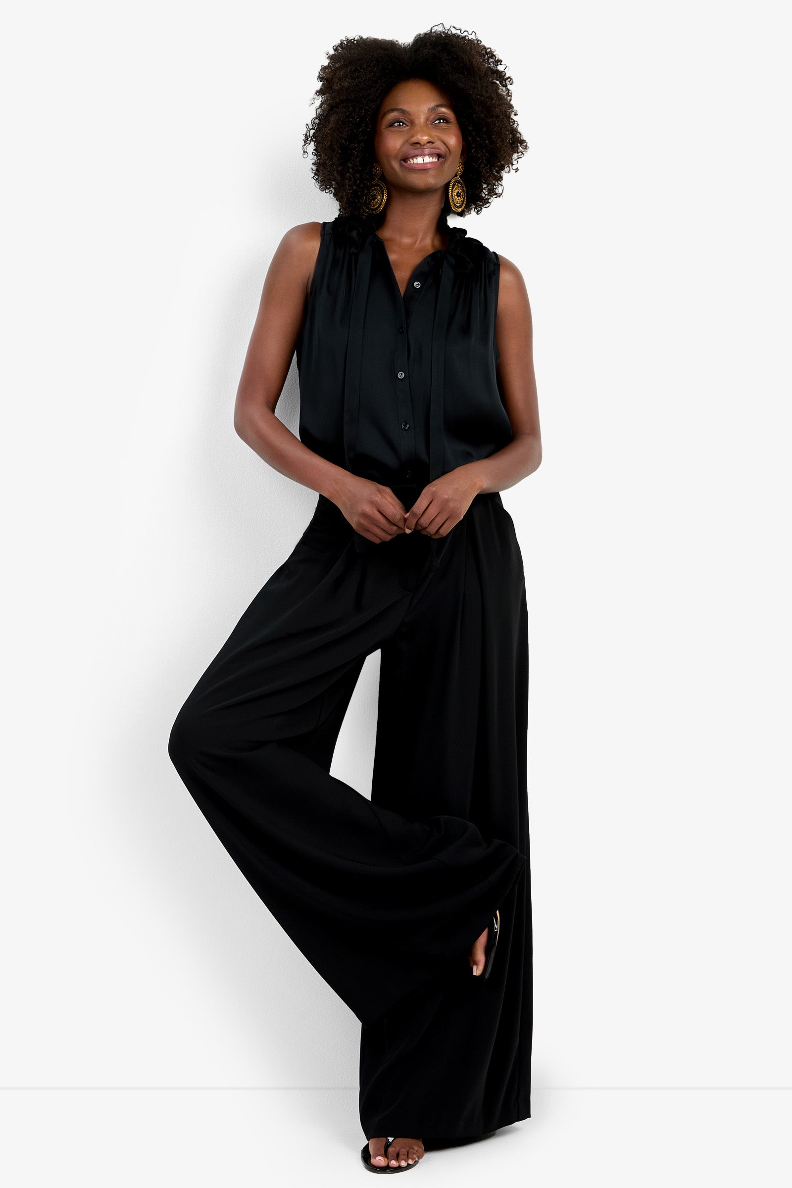 A woman with curly hair smiles against a white background, wearing the Saige Long Pant, a sleeveless black blouse, large round earrings, and black sandals.