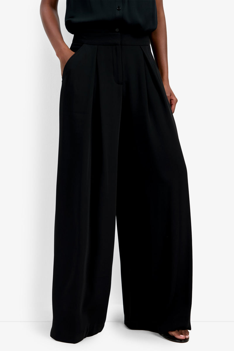 Model wears the Saige Long Pant—high-waisted, wide-leg black trousers with pockets—paired with a black top. The look features a loose, flowing silhouette for an elegant, modern style.