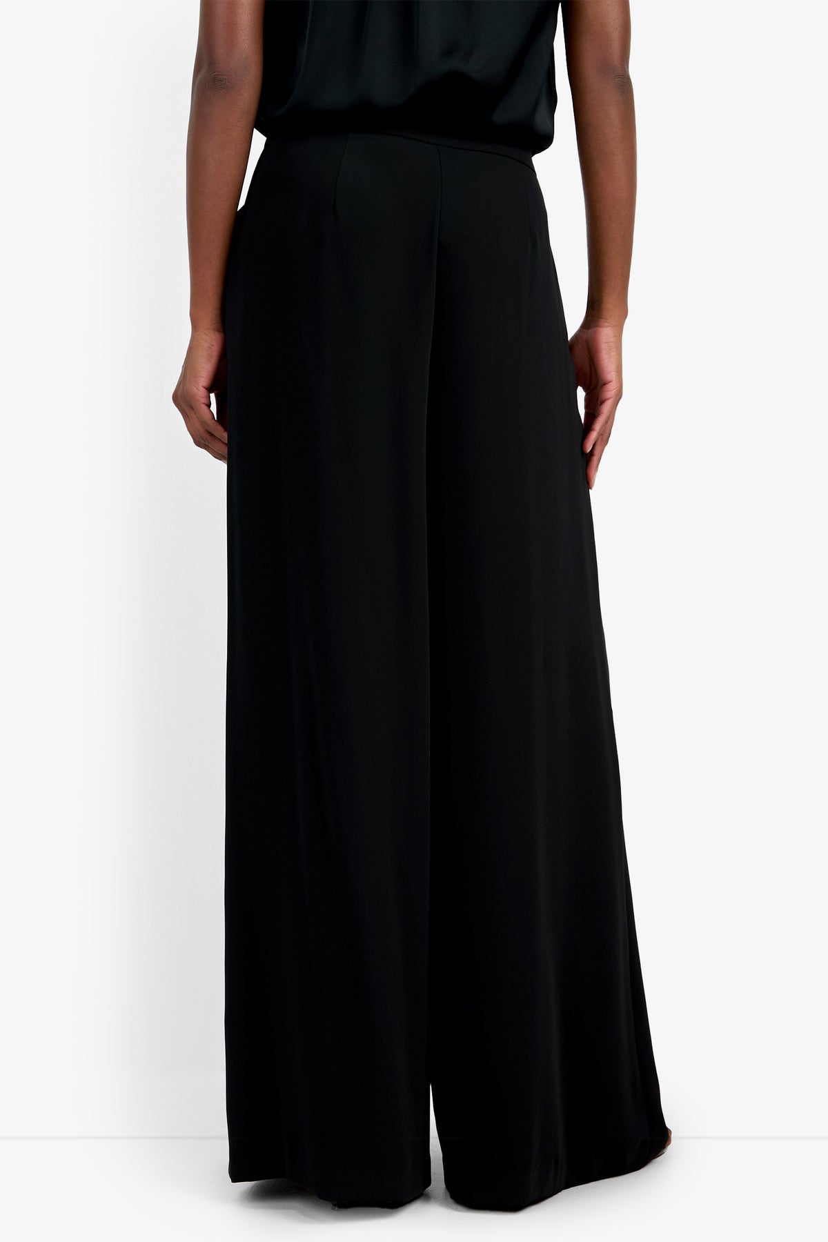 A person stands facing away against a white background, wearing loose-fitting, high-waisted Saige Long Pant in black and a matching black top.