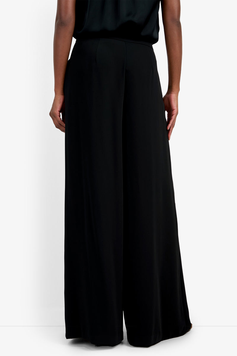 A person stands facing away against a white background, wearing loose-fitting, high-waisted Saige Long Pant in black and a matching black top.