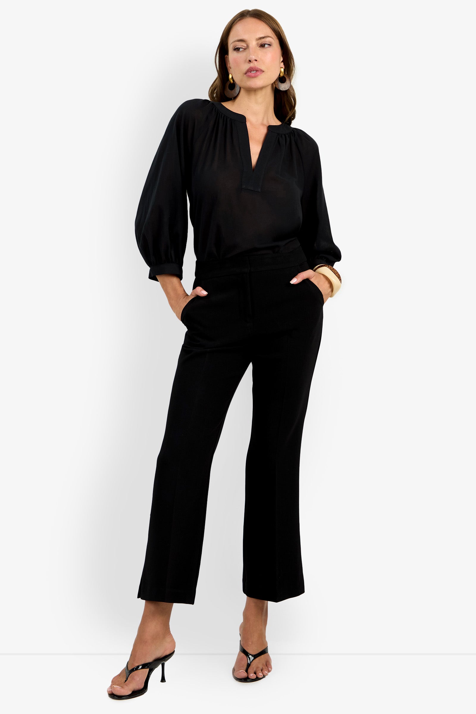 A woman wears the Allisan Pant with a black long-sleeve blouse and black heeled sandals, standing with one hand in her pocket against a white background. She completes the look with gold earrings and a bracelet.