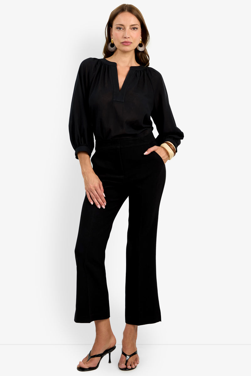 A woman wearing the Allisan Pant, a black long-sleeve blouse, black heeled sandals, and statement earrings stands with one hand in her pocket and the other on her hip against a white background.