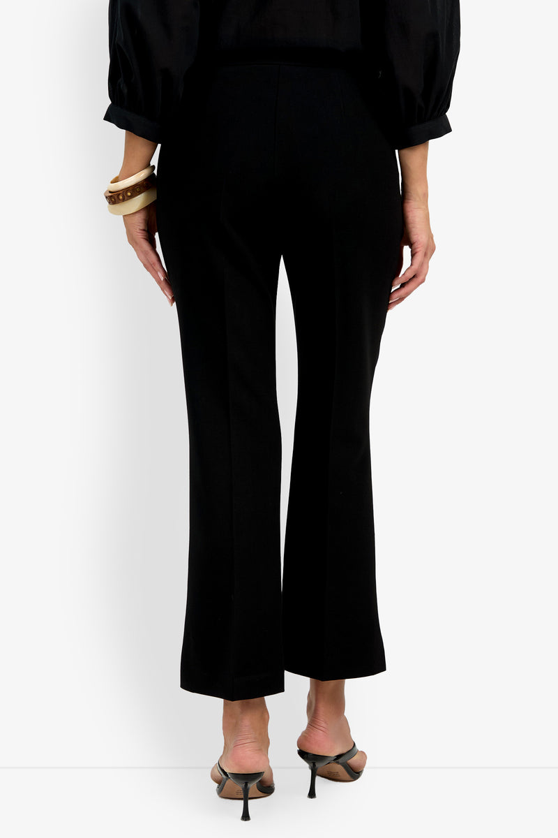 A person stands facing away on a plain white background, wearing the Allisan Pant in black, a black long-sleeve blouse, heeled sandals, and bracelets on their left wrist.