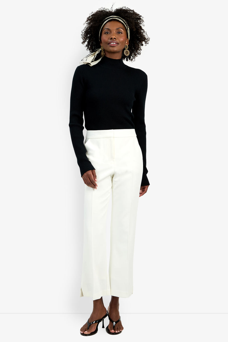 A woman poses against a plain background wearing the Allisan Pant—white, high-waisted, wide-leg trousers—paired with a black turtleneck sweater, black heeled sandals, gold earrings, and a light scarf tied around her head.