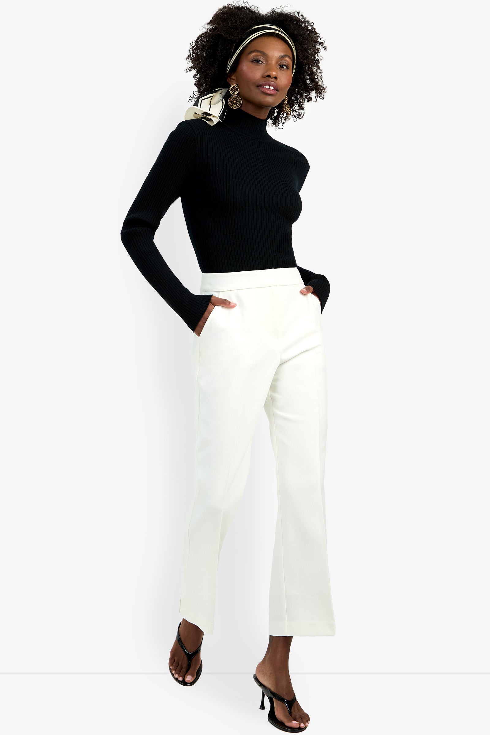 A woman stands confidently facing the camera, hands in her pockets, wearing the Allisan Pant in white with a black turtleneck sweater, black heeled sandals, large hoop earrings, and a patterned headscarf.