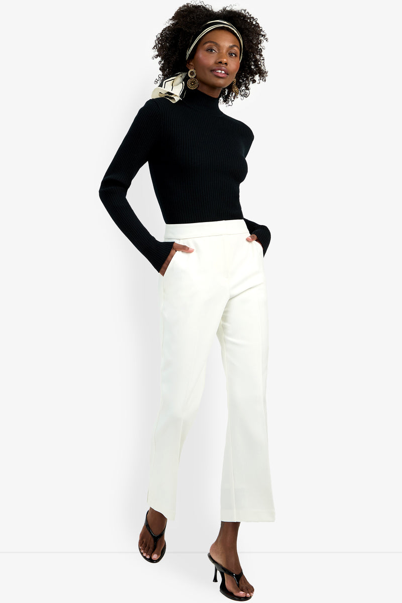 A woman stands confidently facing the camera, hands in her pockets, wearing the Allisan Pant in white with a black turtleneck sweater, black heeled sandals, large hoop earrings, and a patterned headscarf.
