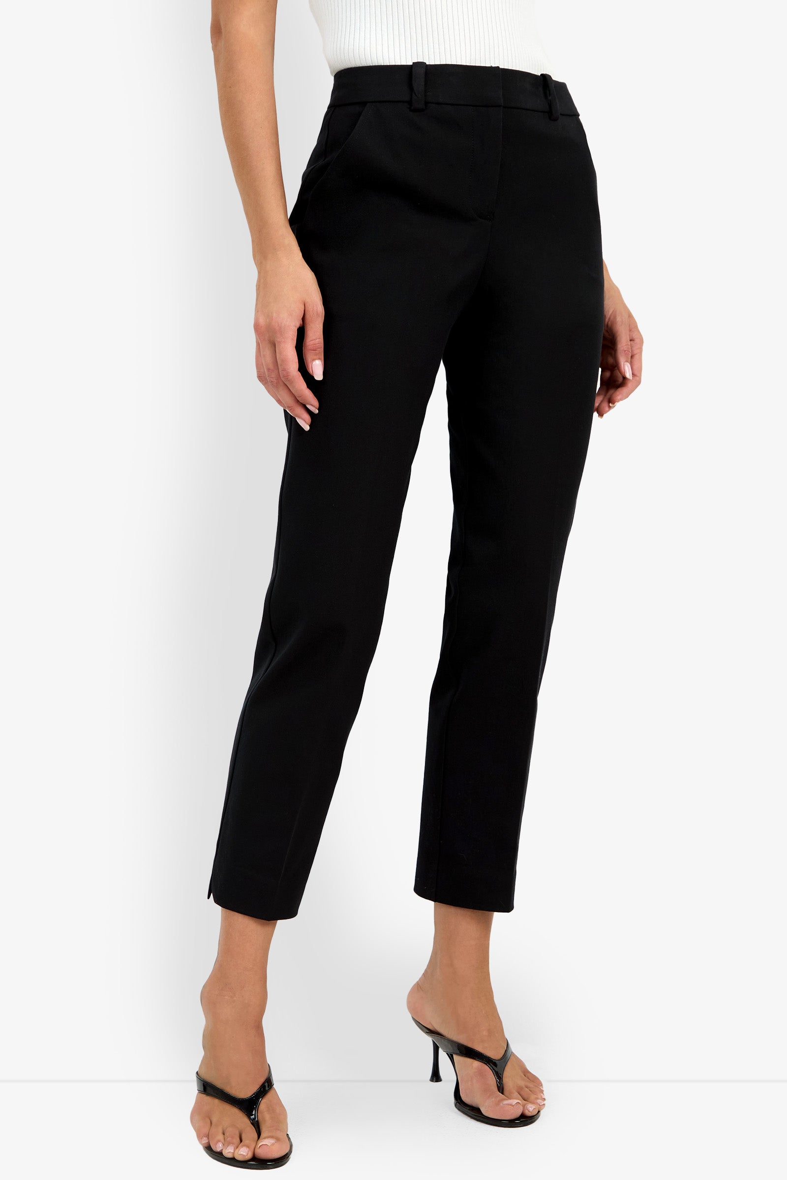 A woman stands against a white background wearing the Audrey Pant in black, ankle-length and tailored, paired with a white sleeveless top and black heeled sandals. Her left hand rests by her side, and her right foot is slightly forward.