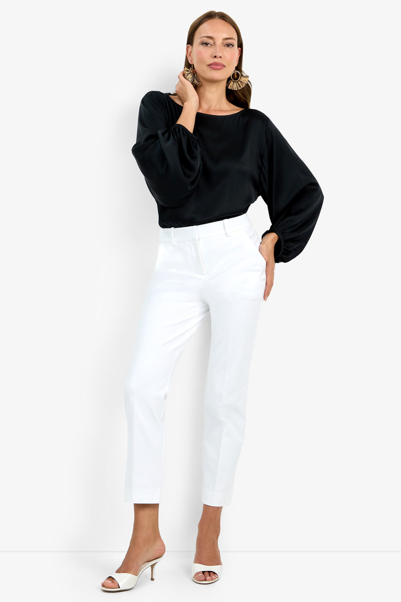 A woman stands confidently in white high-waisted, ankle-length Audrey Pants, a black long-sleeve blouse with wide sleeves, white heeled sandals, and large gold earrings against a plain white background.
