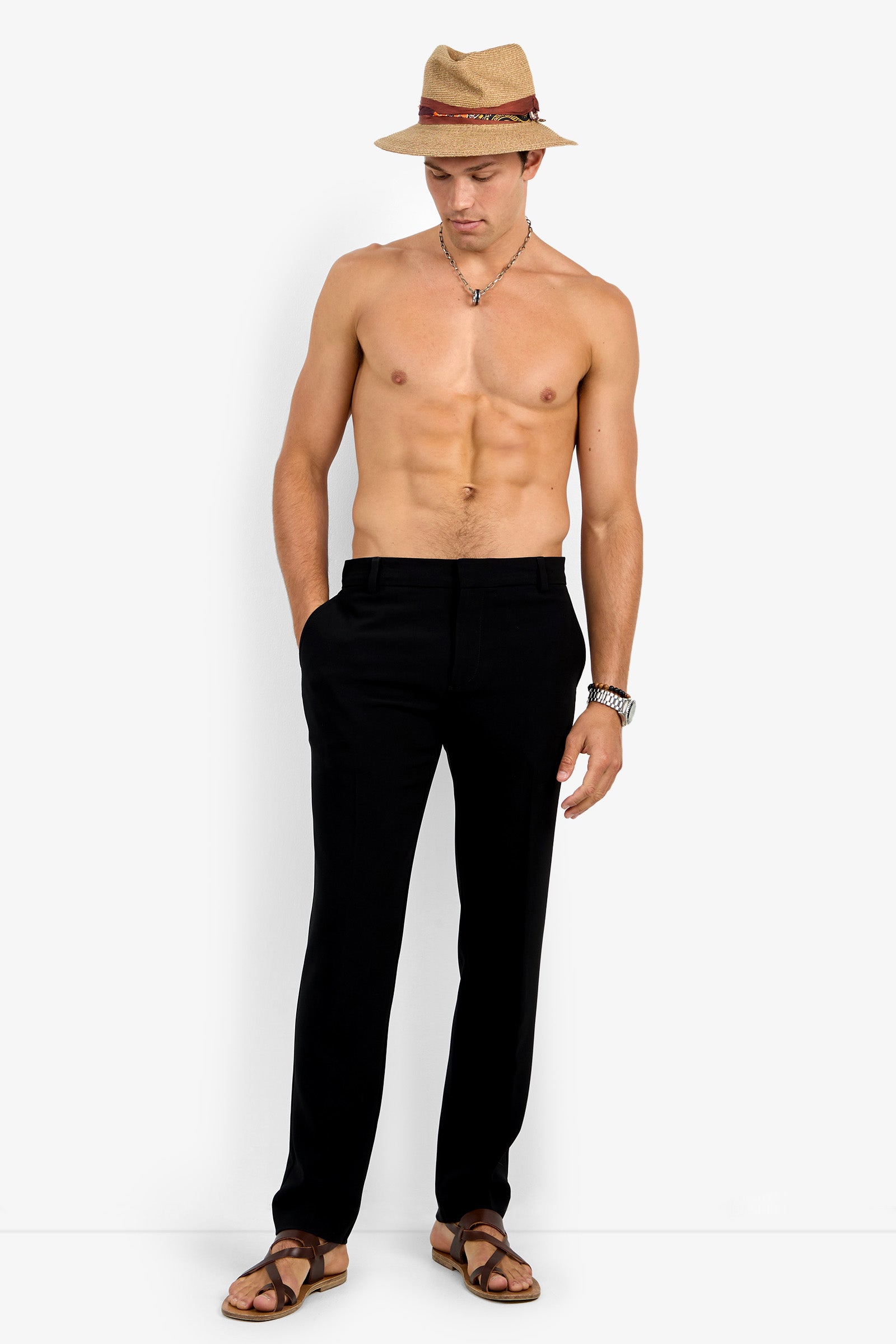 A shirtless man stands with hands in pockets against a plain white background, wearing Clyde Slim Trouser black pants, brown sandals, a straw hat with a red band, a necklace, and a wristwatch.