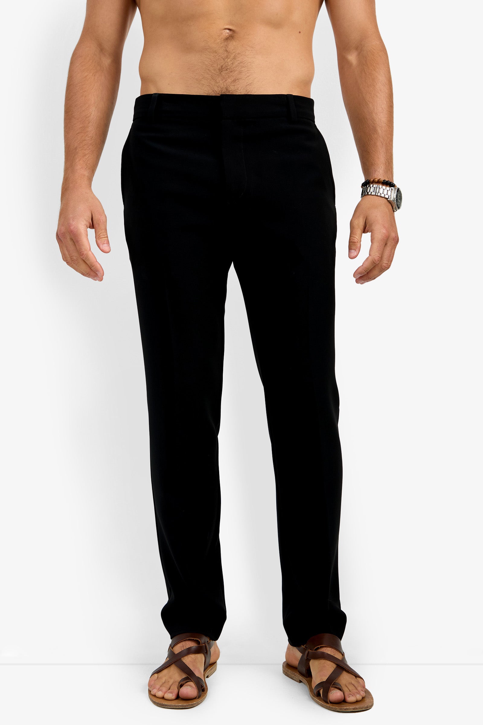 A man wears the Clyde Slim Trouser in black with brown sandals and a wristwatch, standing against a plain white background. The image is cropped from his shoulders down, focusing on the pants.