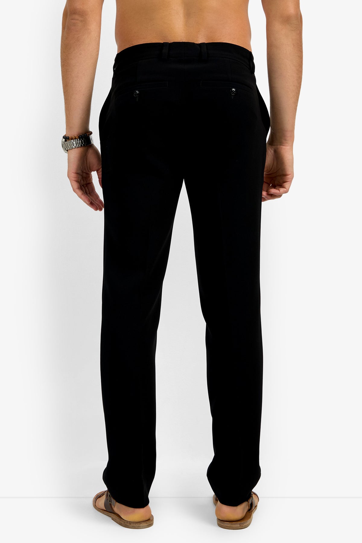 A man with his back to the camera wears Clyde Slim Trouser black dress pants, brown sandals, a wristwatch, and no shirt, set against a plain white background.