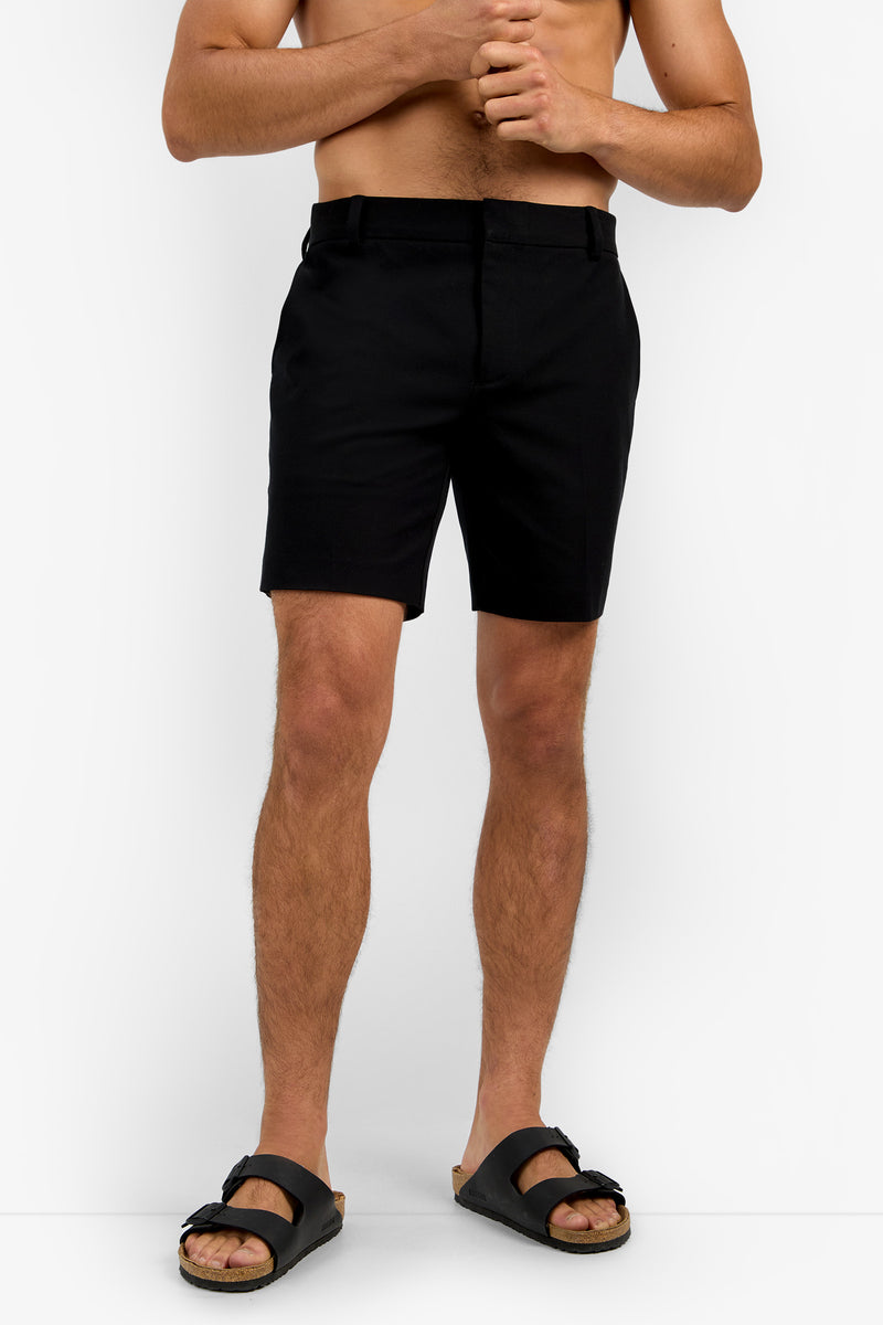 A shirtless man models the Lawrence Short in black with matching sandals, standing against a white background. His hands are clasped near his chest, and his face is not shown.