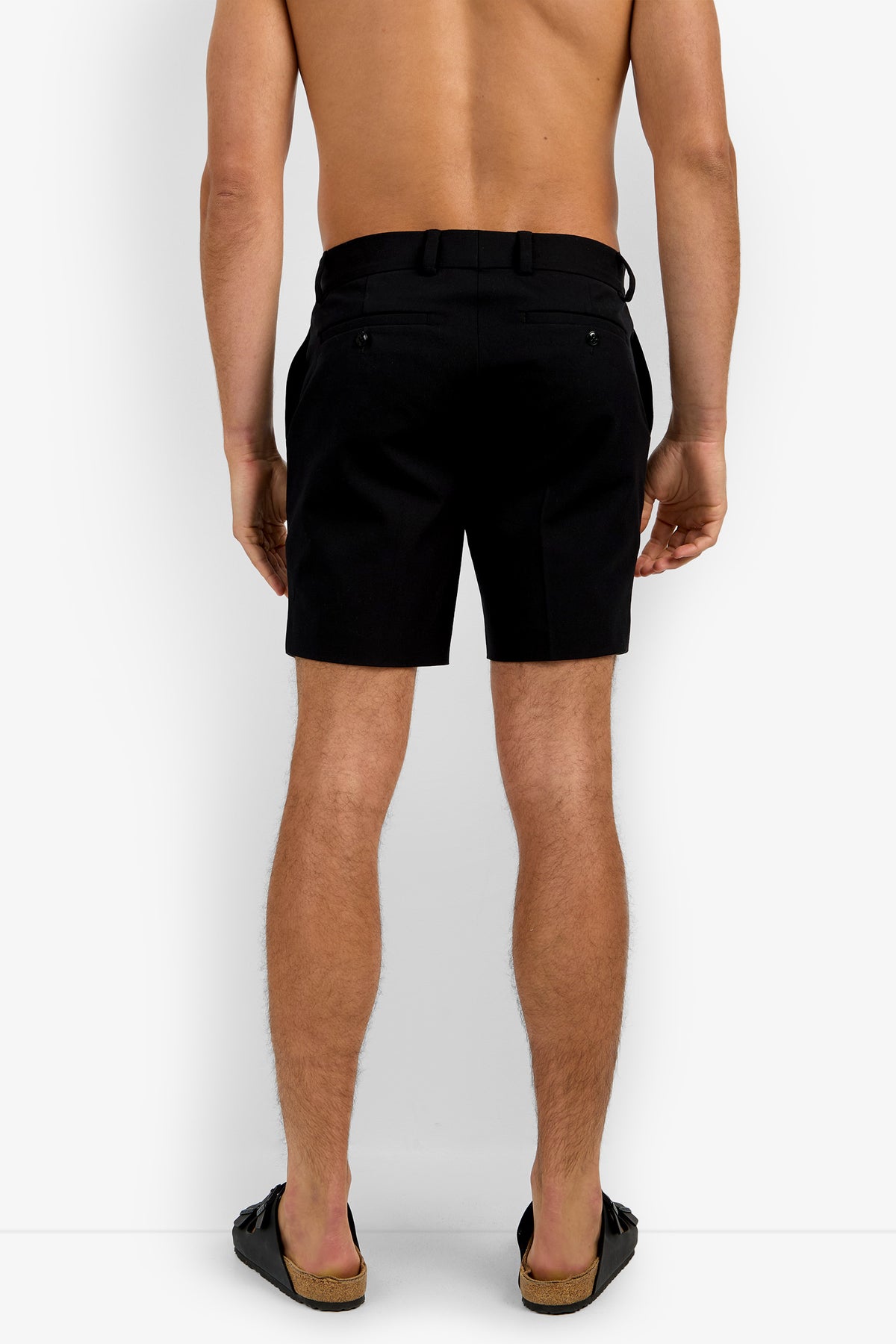 A shirtless man stands facing away, wearing the Lawrence Short in black and black slide sandals against a plain white background.