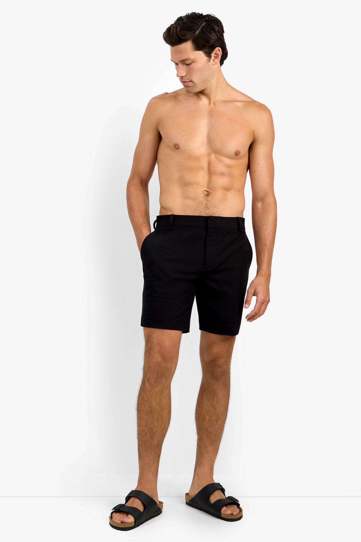 A shirtless man stands against a white background, wearing the Lawrence Short in black and black sandals. He has his left hand in his pocket and looks down toward his right side.