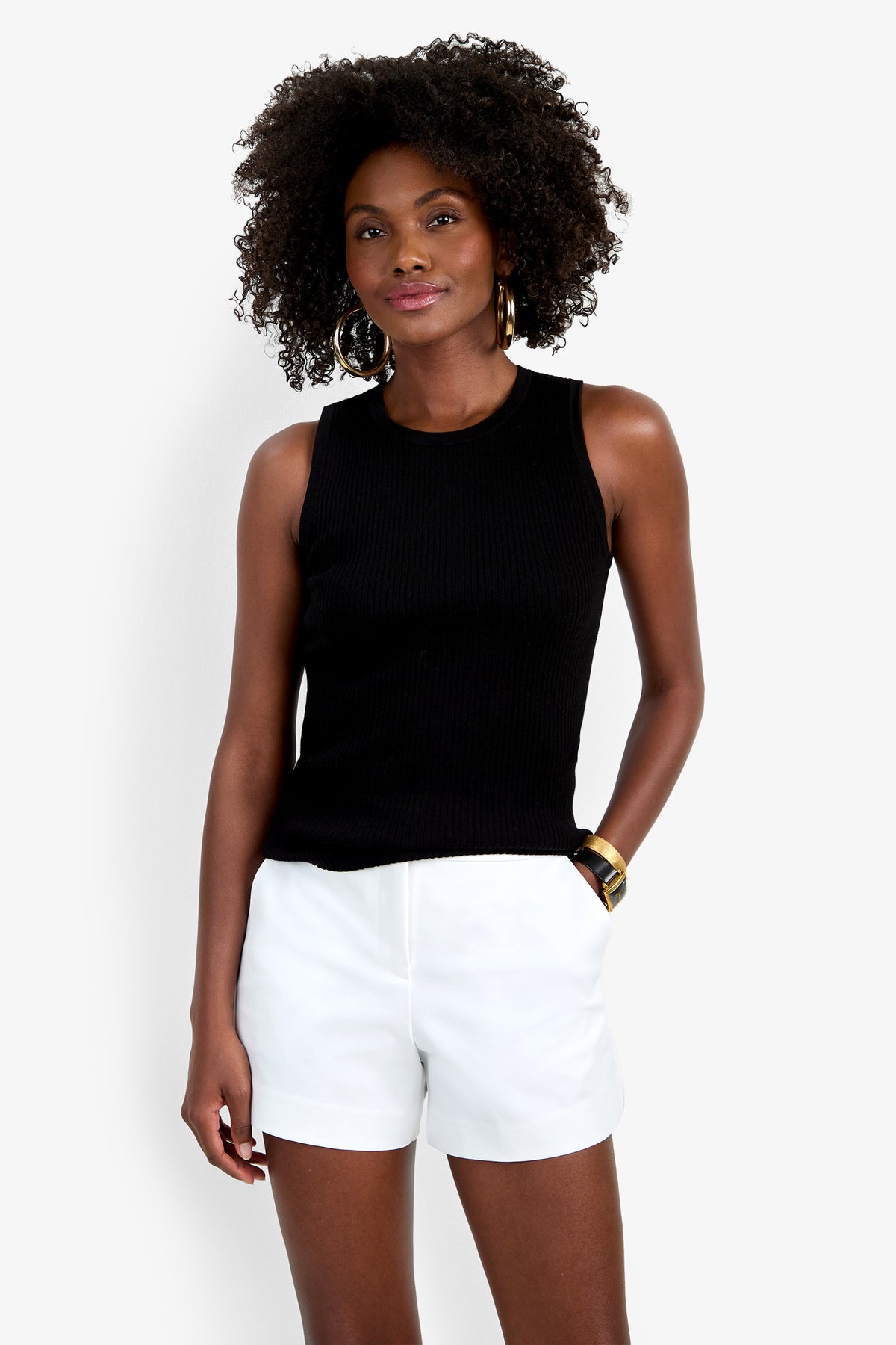 A woman with curly hair smiles softly against a plain white background, wearing the sleeveless black Hampton Crewneck Top, white shorts, gold hoop earrings, and a gold bracelet.