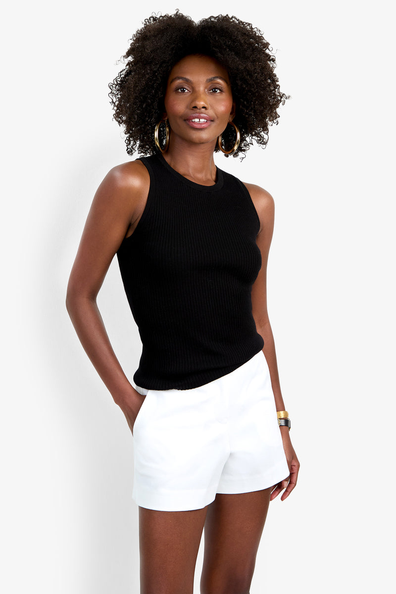 A woman with curly hair wears the Hampton Crewneck Top in black, paired with white shorts and gold hoop earrings, standing against a plain white background with one hand in her pocket.