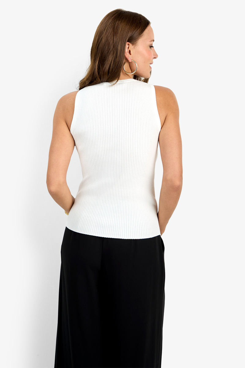 A woman with long brown hair, wearing large hoop earrings and the white ribbed sleeveless Hampton Crewneck Top with black pants, stands facing away from the camera against a plain white background.