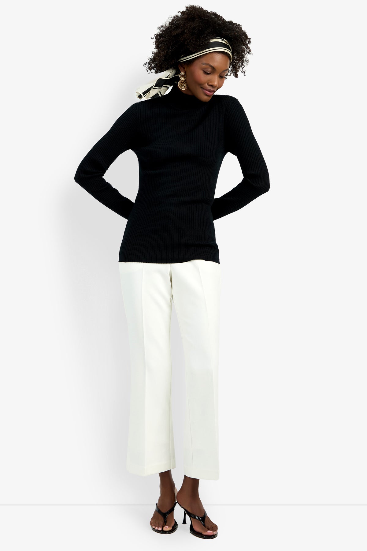 A woman stands against a white background wearing the black Hempstead Mock Neck Top, white wide-leg pants, black heeled sandals, gold earrings, and a black-and-white headscarf.