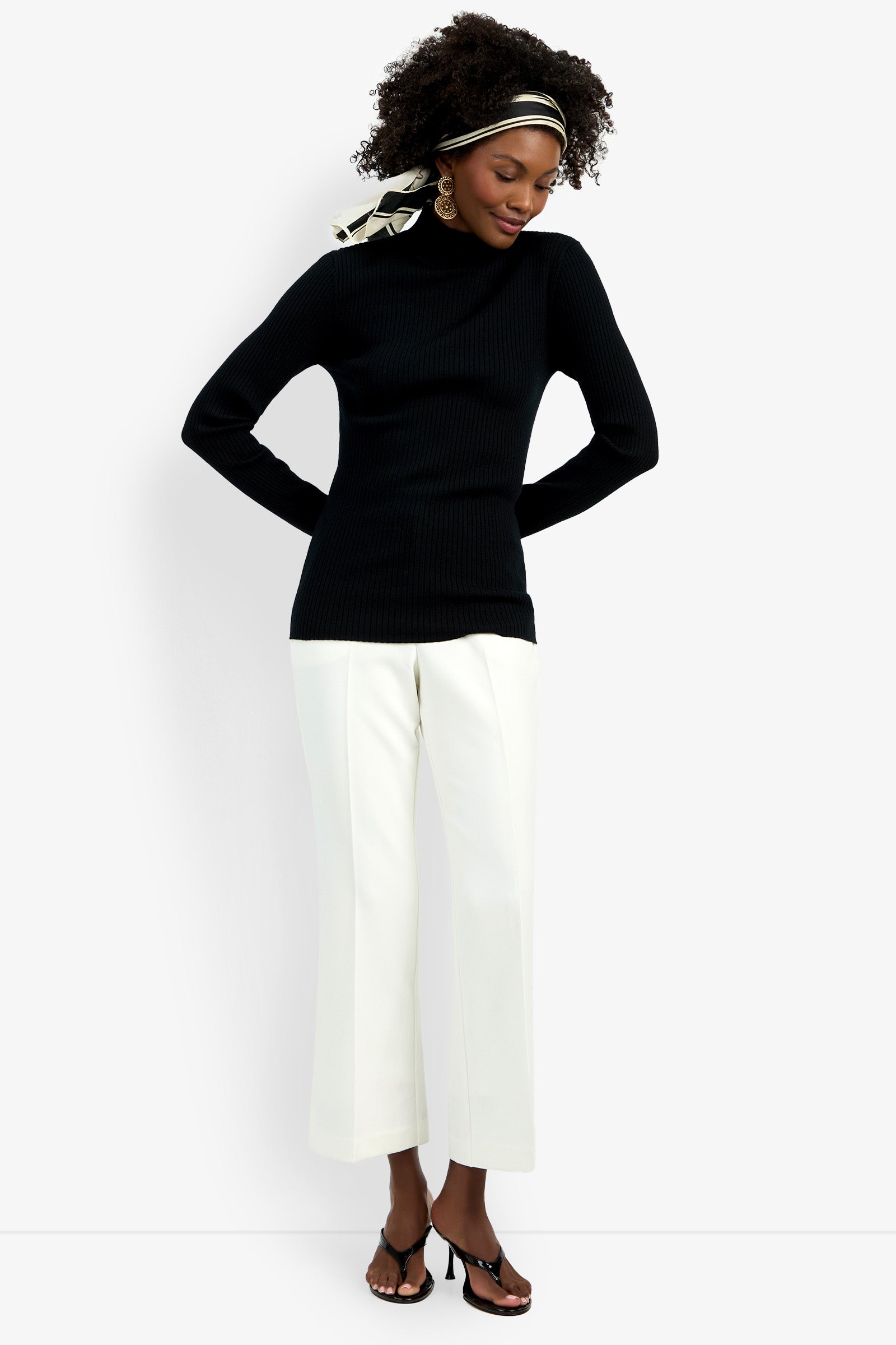 A woman stands against a white background wearing the black Hempstead Mock Neck Top, white wide-leg pants, black heeled sandals, gold earrings, and a black-and-white headscarf.