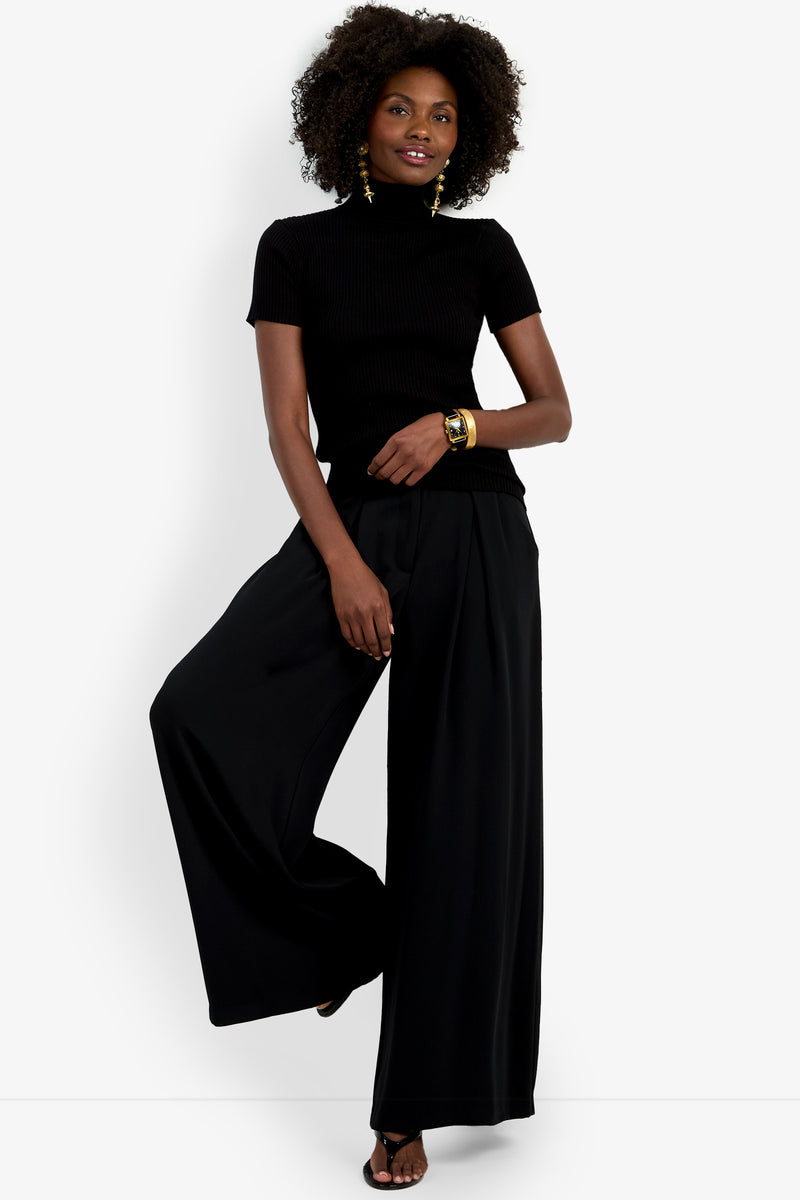A woman with curly hair poses against a white background, wearing the Armeni Mock Neck Top in black with wide-leg black pants, gold earrings, a gold bracelet, and black sandals; she lifts one leg.