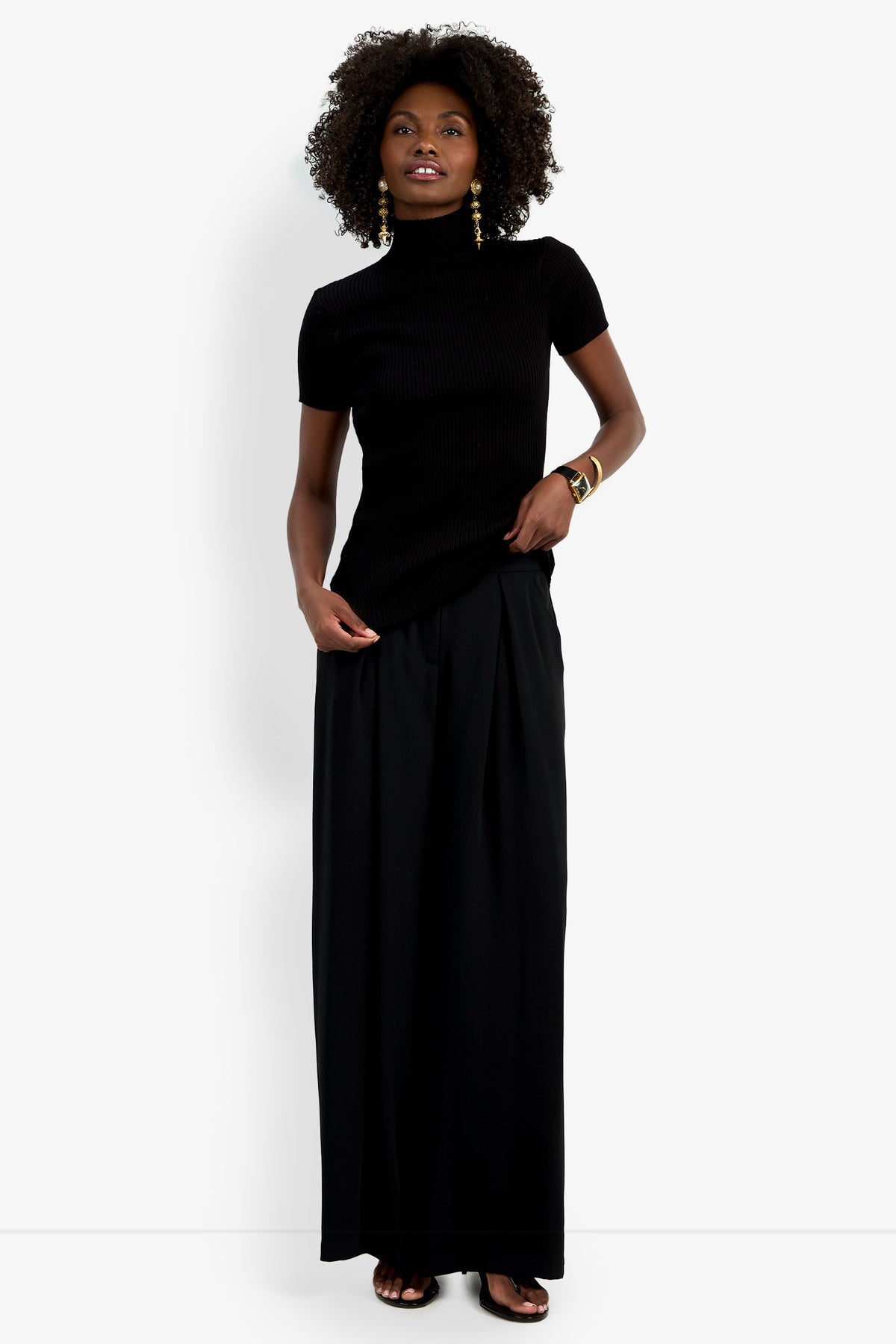 A woman with curly hair smiles slightly against a white background, wearing the Armeni Mock Neck Top, long black wide-leg pants, gold earrings, bangles, and sandals.