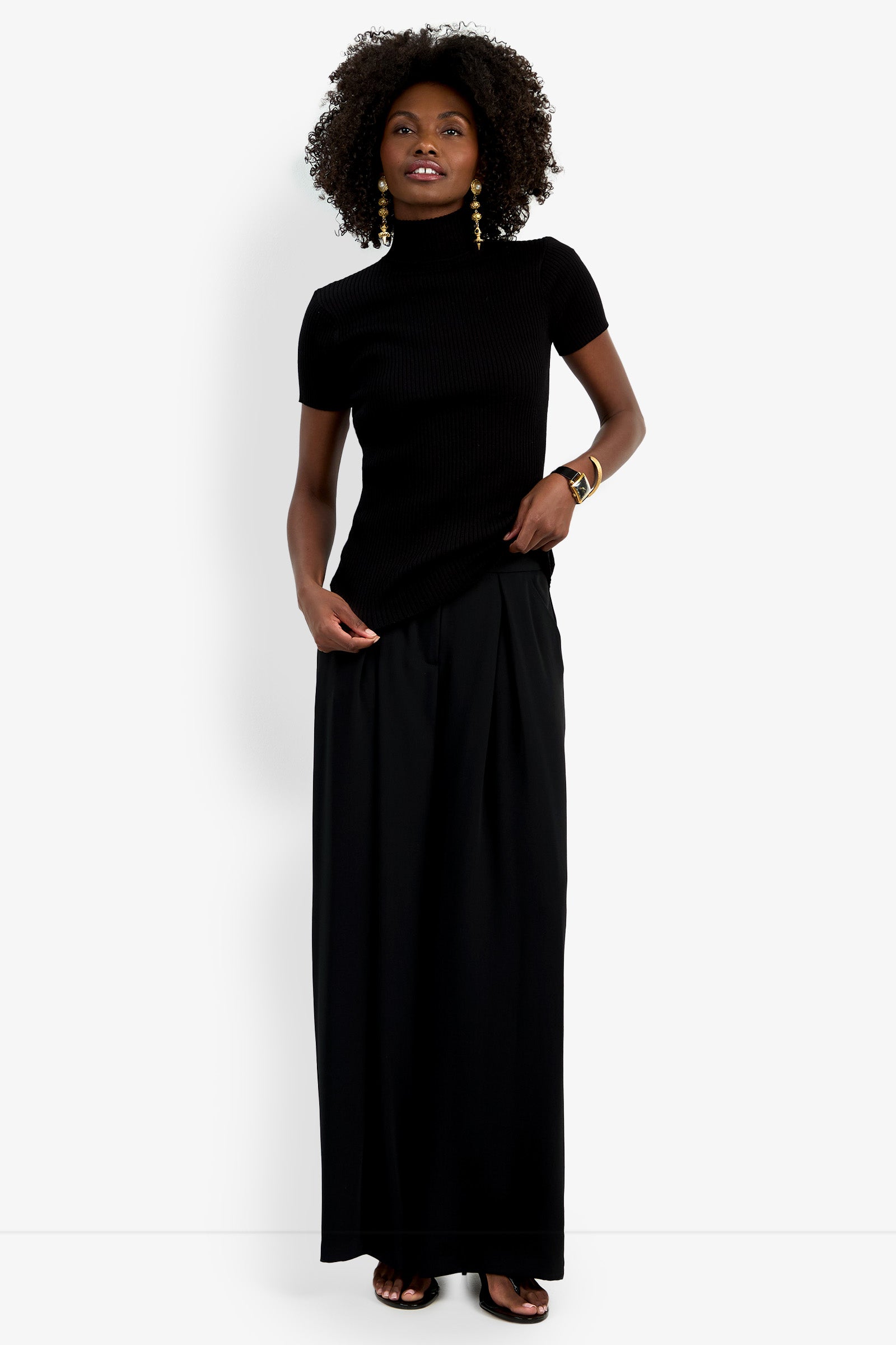 A woman with curly hair smiles slightly against a white background, wearing the Armeni Mock Neck Top, long black wide-leg pants, gold earrings, bangles, and sandals.