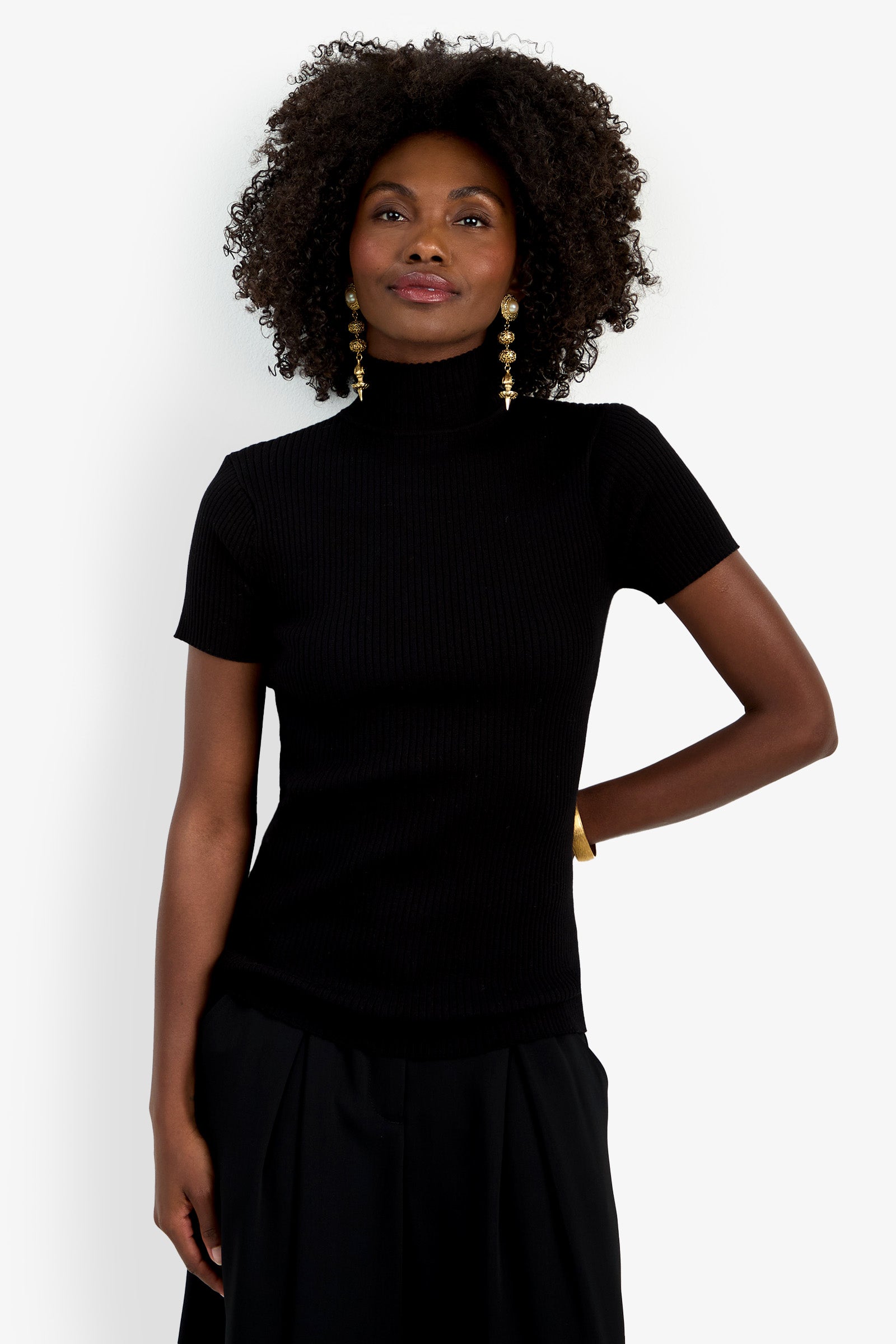 A woman with curly hair smiles gently against a plain white background, wearing the black Armeni Mock Neck Top, long gold statement earrings, and a gold bracelet.