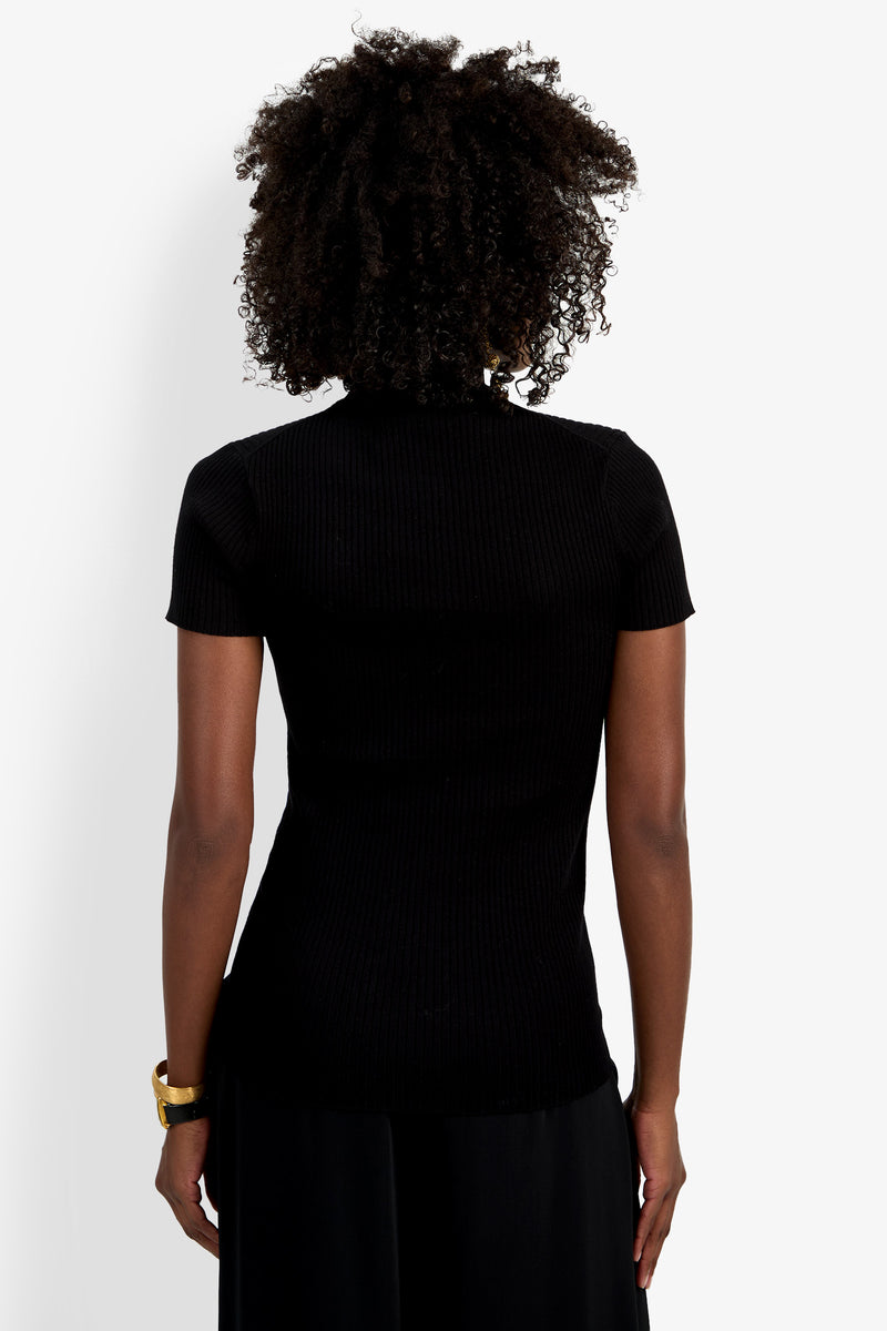 A person with curly hair, wearing the Armeni Mock Neck Top and a gold bracelet, stands facing away from the camera against a plain white background.