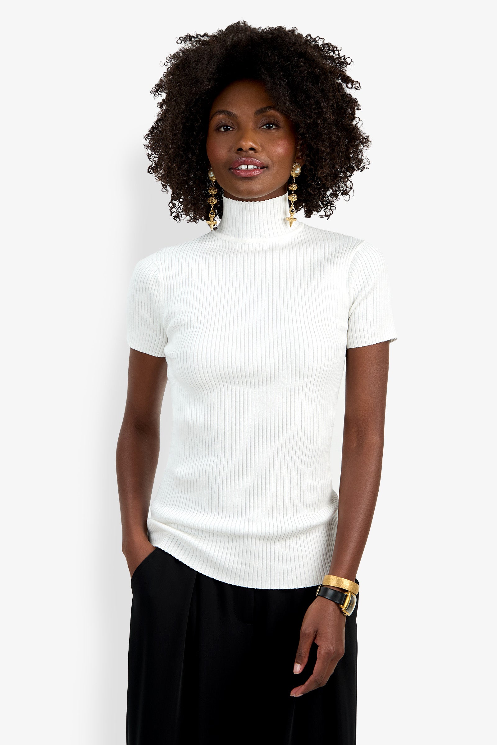 A woman with curly hair wears the Armeni Mock Neck Top—a white ribbed short-sleeve turtleneck—paired with black pants, gold hoop earrings, and a gold bracelet, standing against a plain white background with one hand in her pocket.