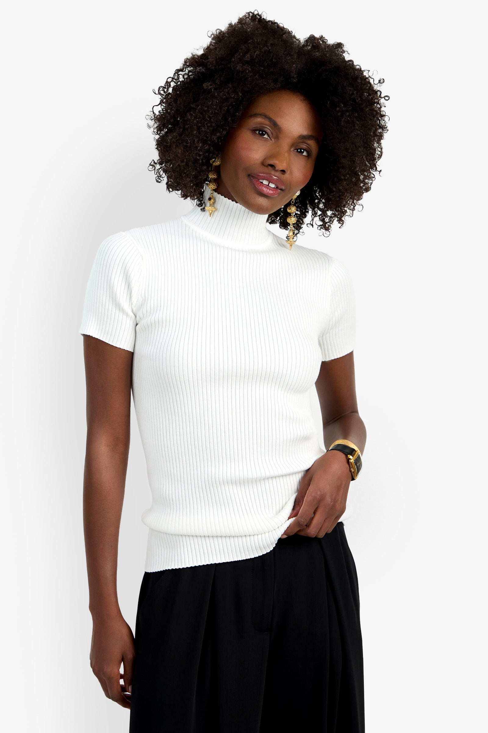 A woman with curly hair wears the Armeni Mock Neck Top, a white ribbed short-sleeve turtleneck, paired with black pants and accessorized with dangling earrings and a watch. She stands against a white background, smiling slightly.
