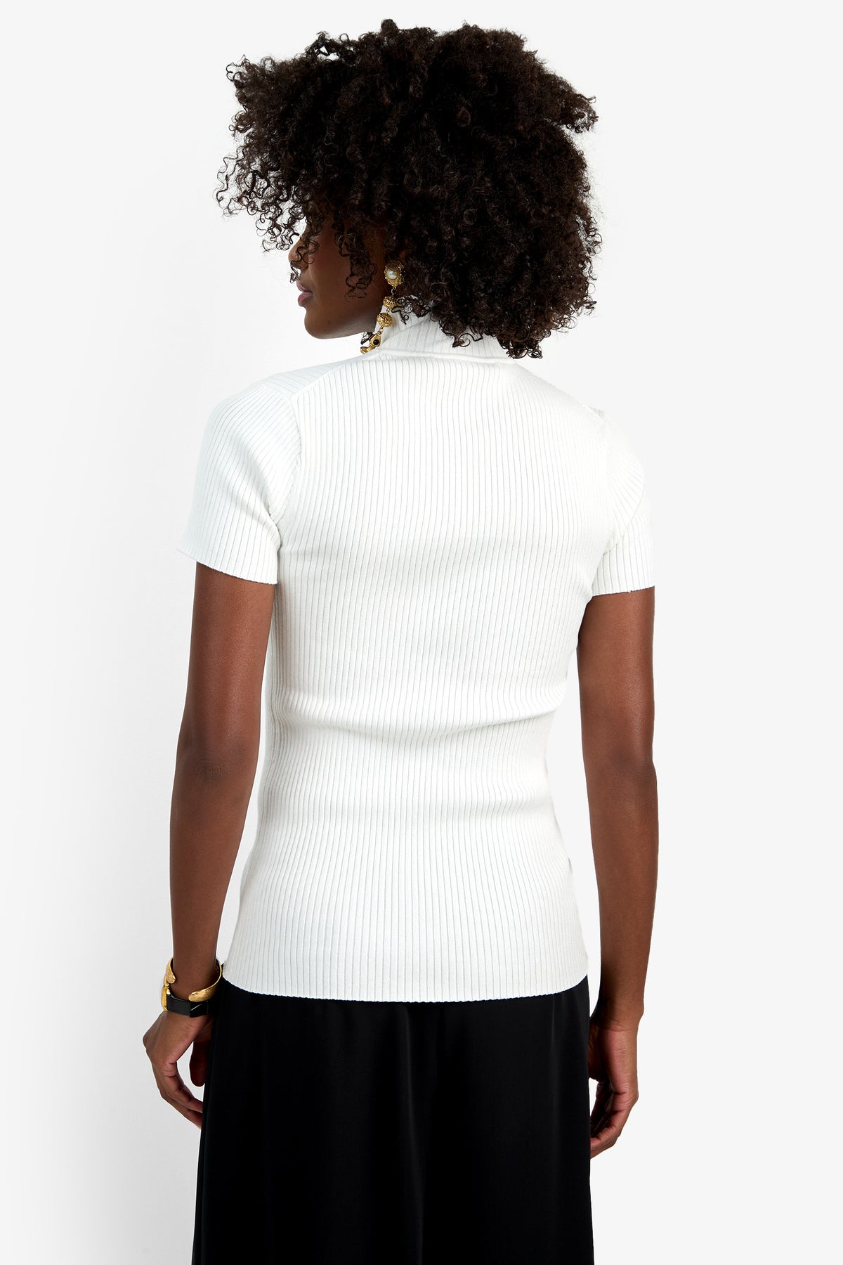 A person with curly dark hair stands with their back to the camera, wearing the Armeni Mock Neck Top in white and black pants, set against a plain white background.