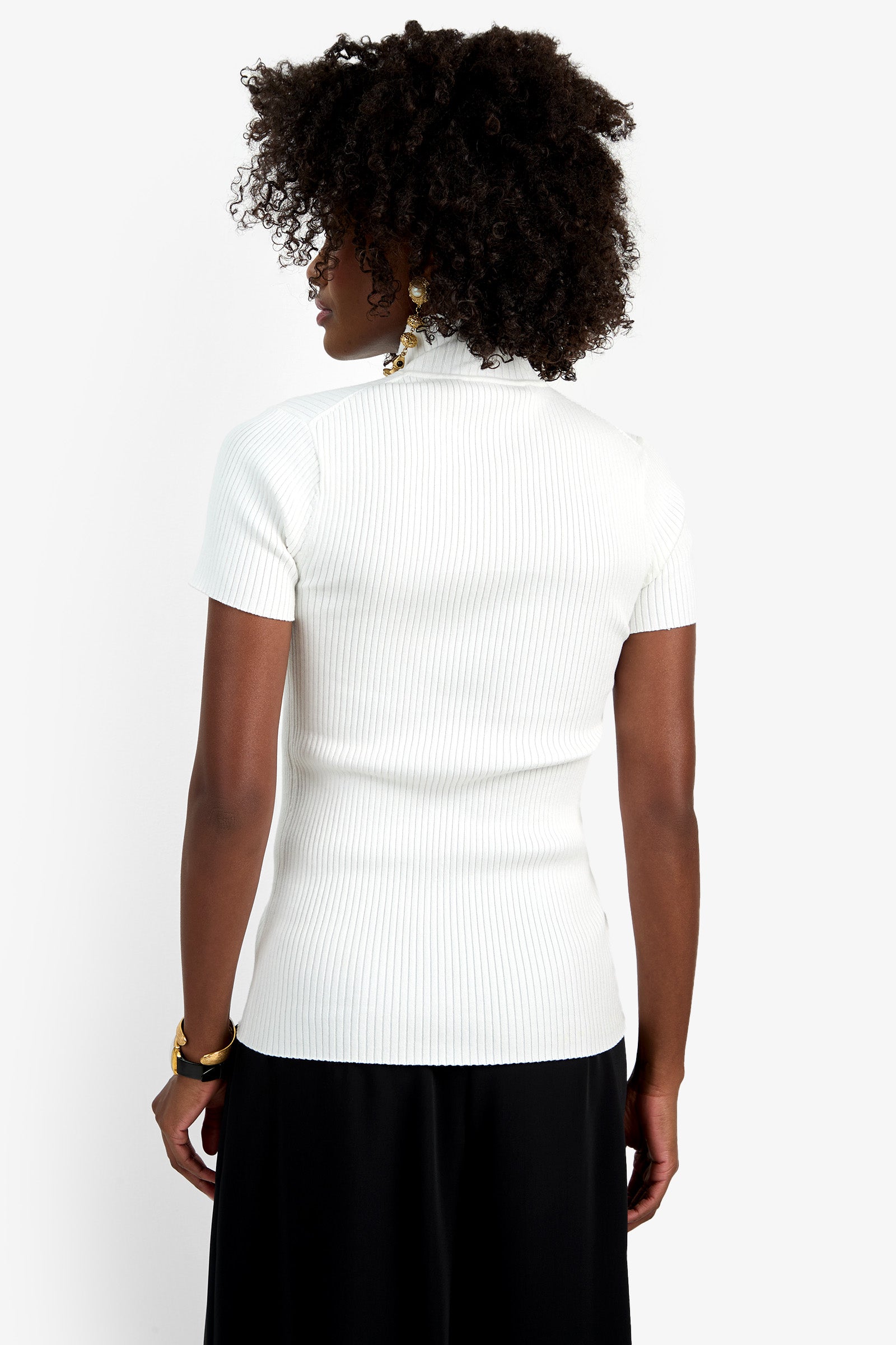 A person with curly dark hair stands with their back to the camera, wearing the Armeni Mock Neck Top in white and black pants, set against a plain white background.