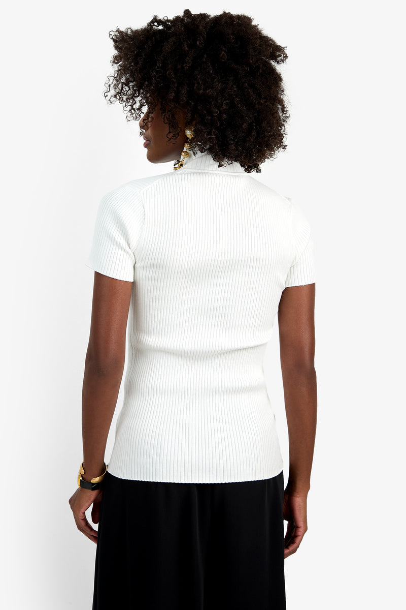 A person with curly dark hair stands with their back to the camera, wearing the Armeni Mock Neck Top in white and black pants, set against a plain white background.
