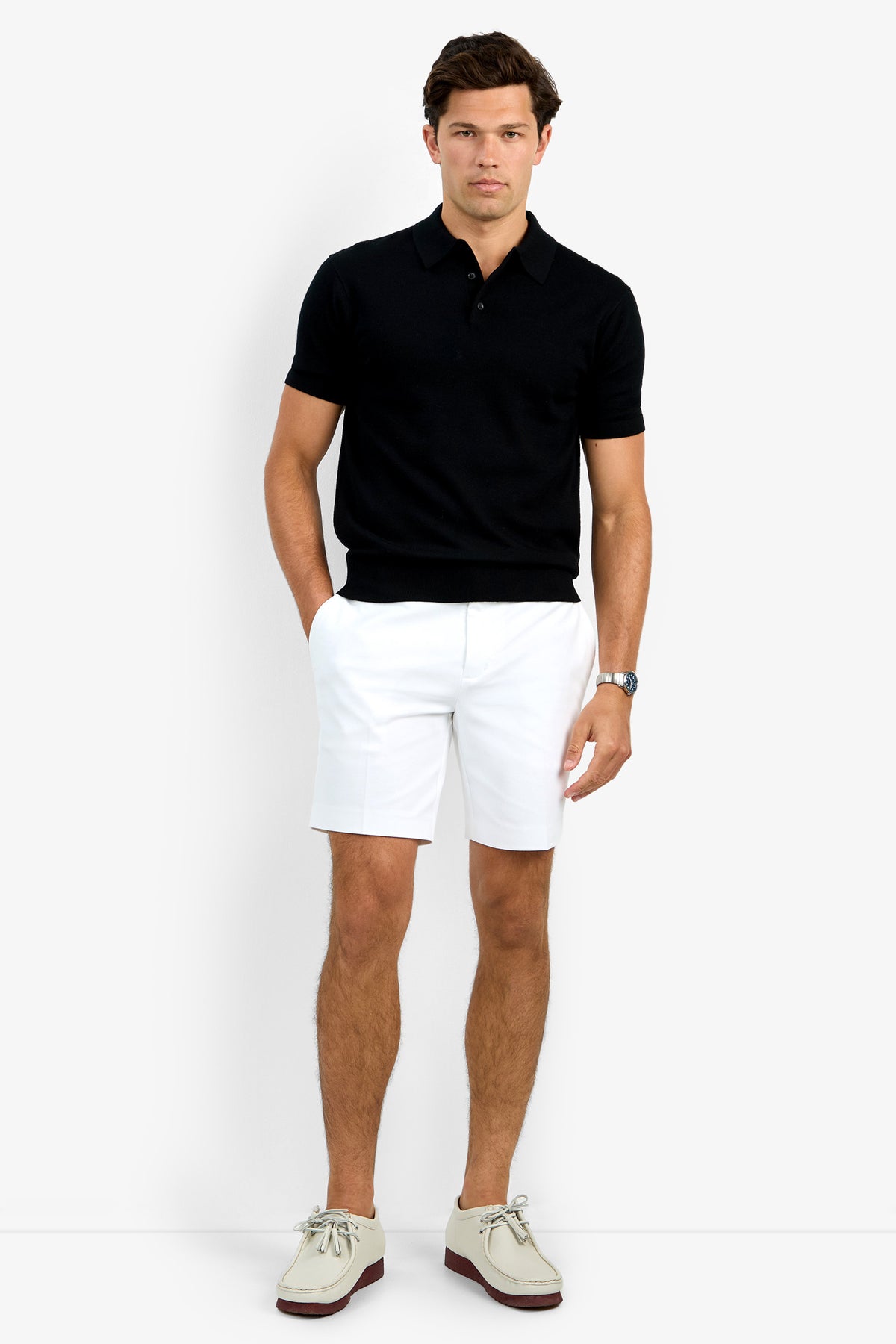 A man stands against a plain white background wearing the Grant Short Sleeve Polo in black, paired with white shorts, beige boat shoes, and a wristwatch, with his hands in his pockets.