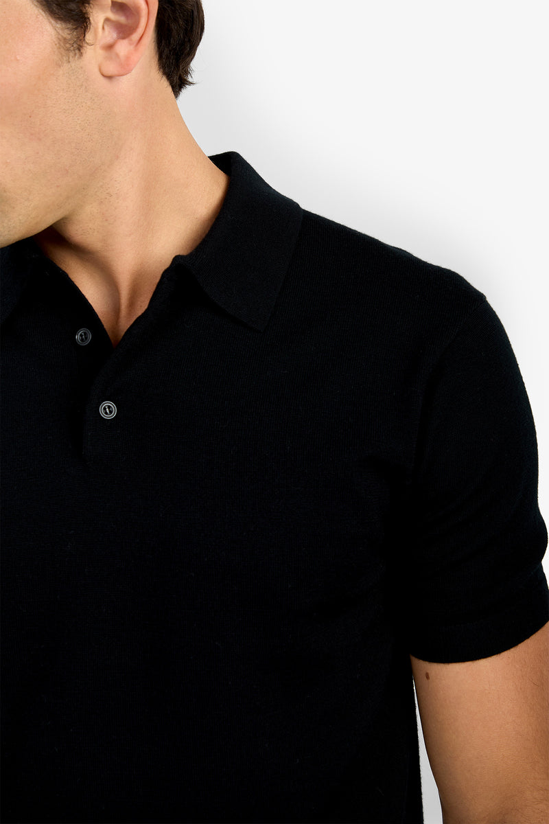 A man models the Grant Short Sleeve Polo—black, with a collar and buttons—shown from shoulders to mid-torso against a plain white background. His face is partially out of frame.