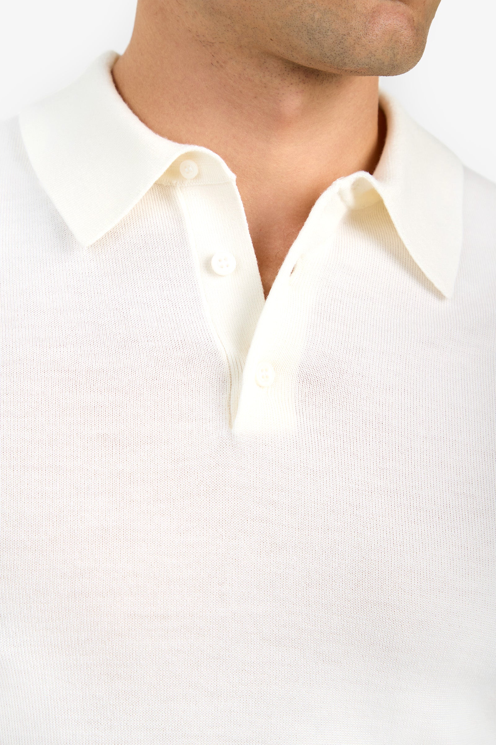 Close-up of a person wearing the Grant Short Sleeve Polo, an off-white knit shirt with a collar and three buttons, showing the upper chest and neck. The background is plain and light-colored.
