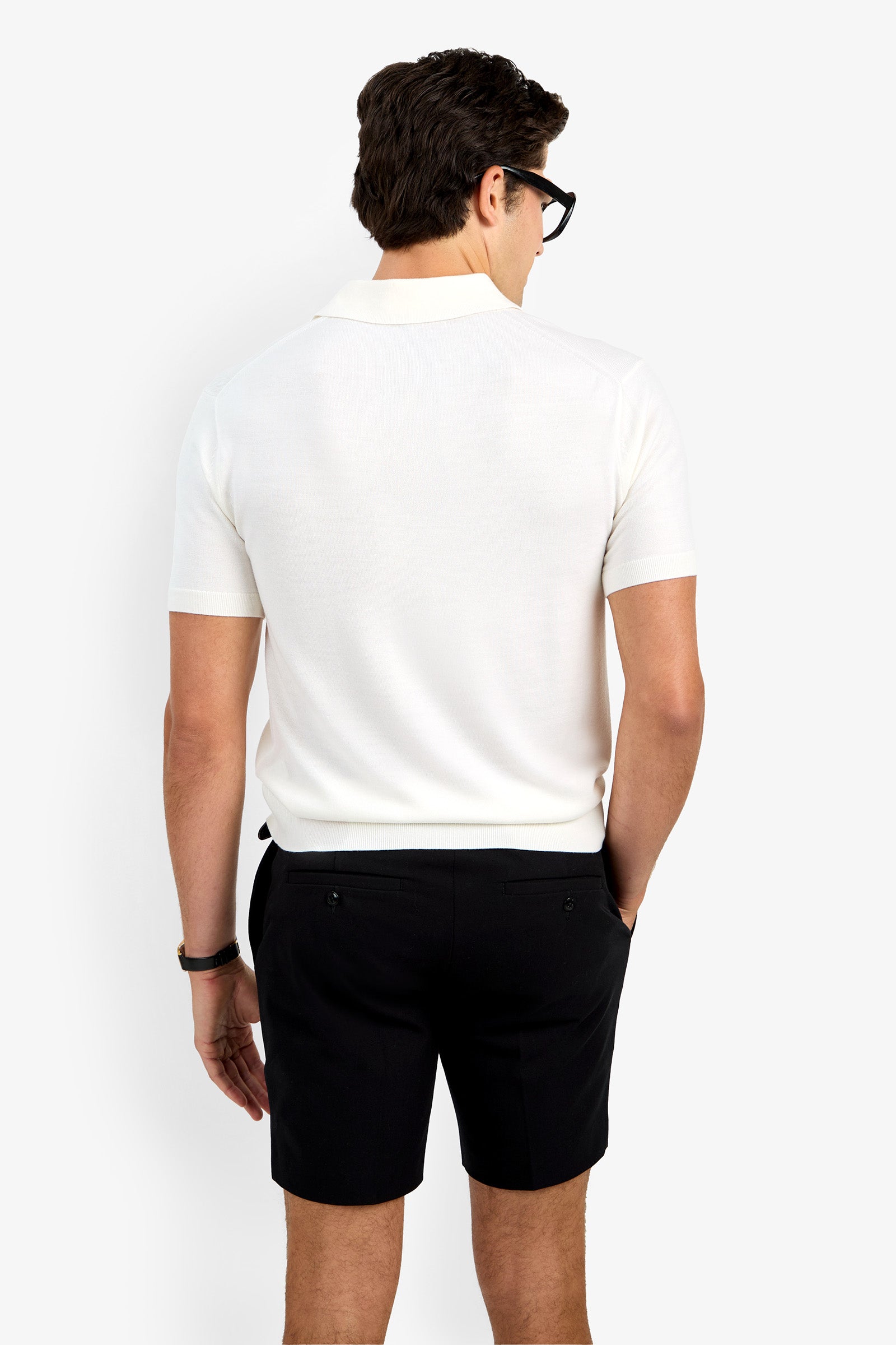 A man with short brown hair, black glasses, and black shorts wears the Grant Short Sleeve Polo in white, standing with his back to the camera against a plain white background.