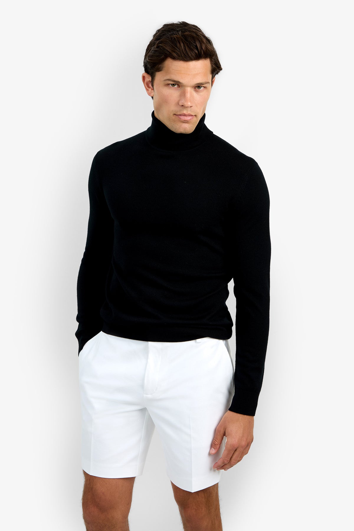 A man stands against a plain white background, wearing the Roy Long Sleeve Turtleneck in black and white knee-length shorts, with one hand in his pocket and a neutral expression.