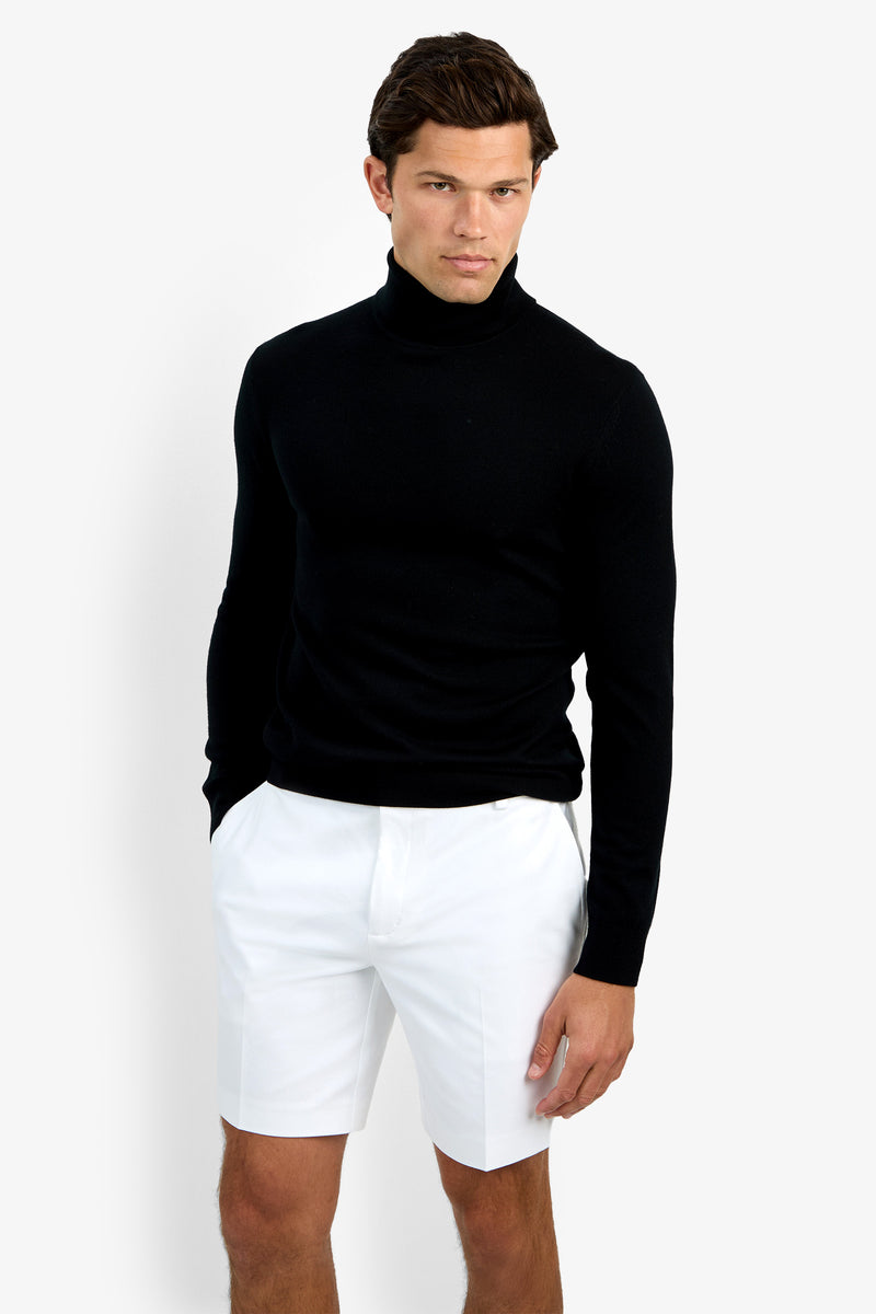 A man confidently poses against a white background, wearing the Roy Long Sleeve Turtleneck in black and white shorts, his left hand by his side and right hand slightly in his pocket.