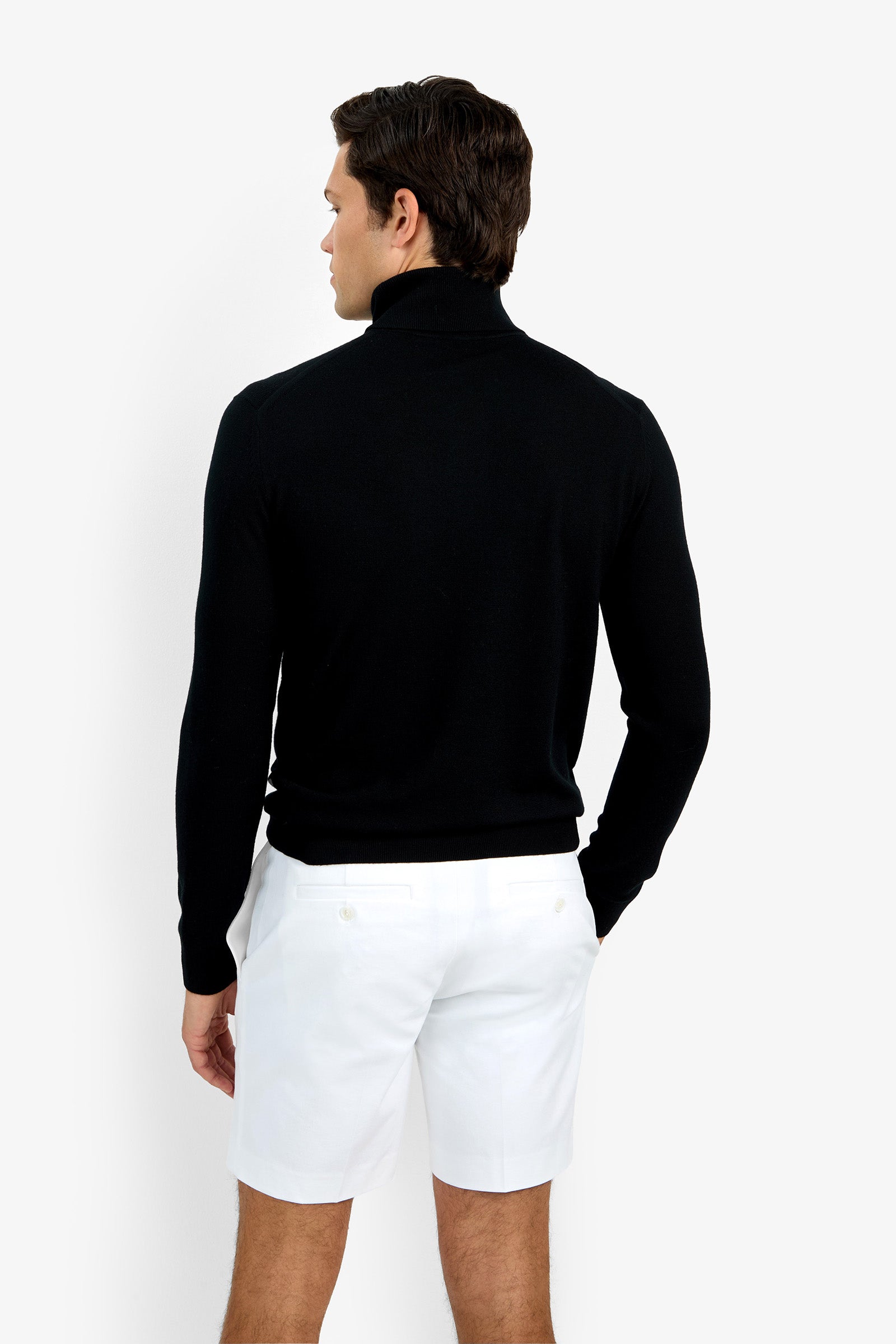 A man faces away from the camera, modeling the Roy Long Sleeve Turtleneck in black with white shorts against a plain white background.